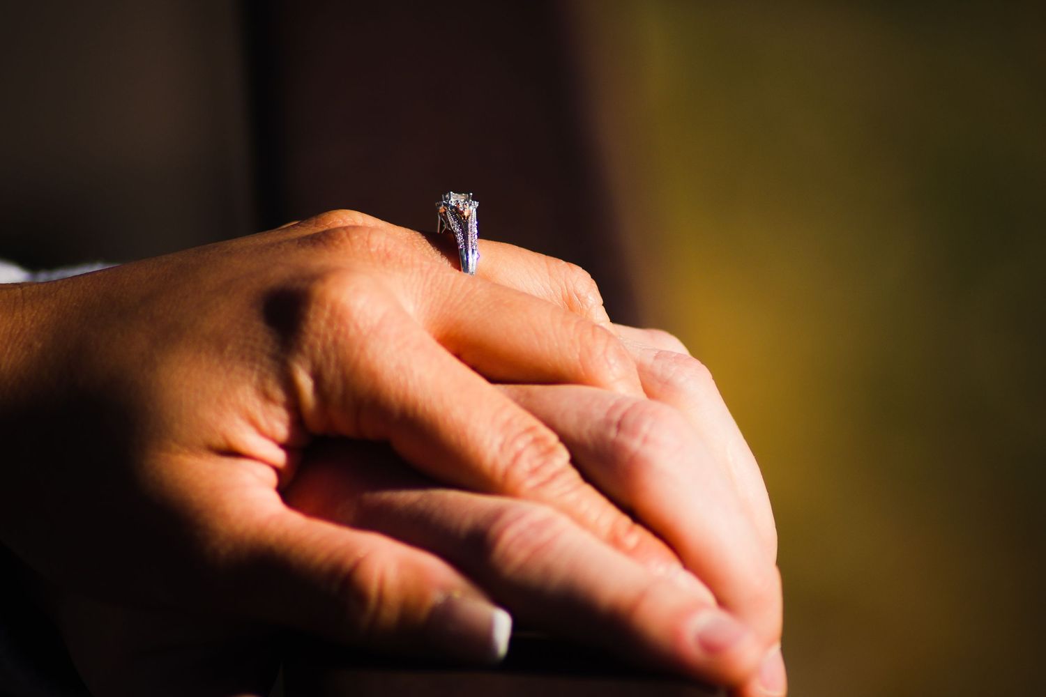 Close-up of clasped hands wearing a sparkling diamond engagement ring in warm light.