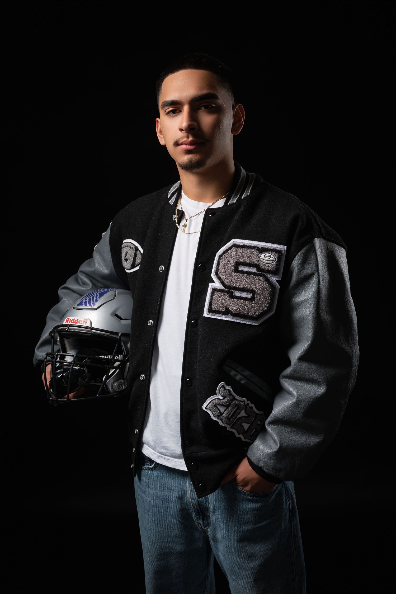 A young man in a varsity letterman jacket holds a football helmet against a black background.