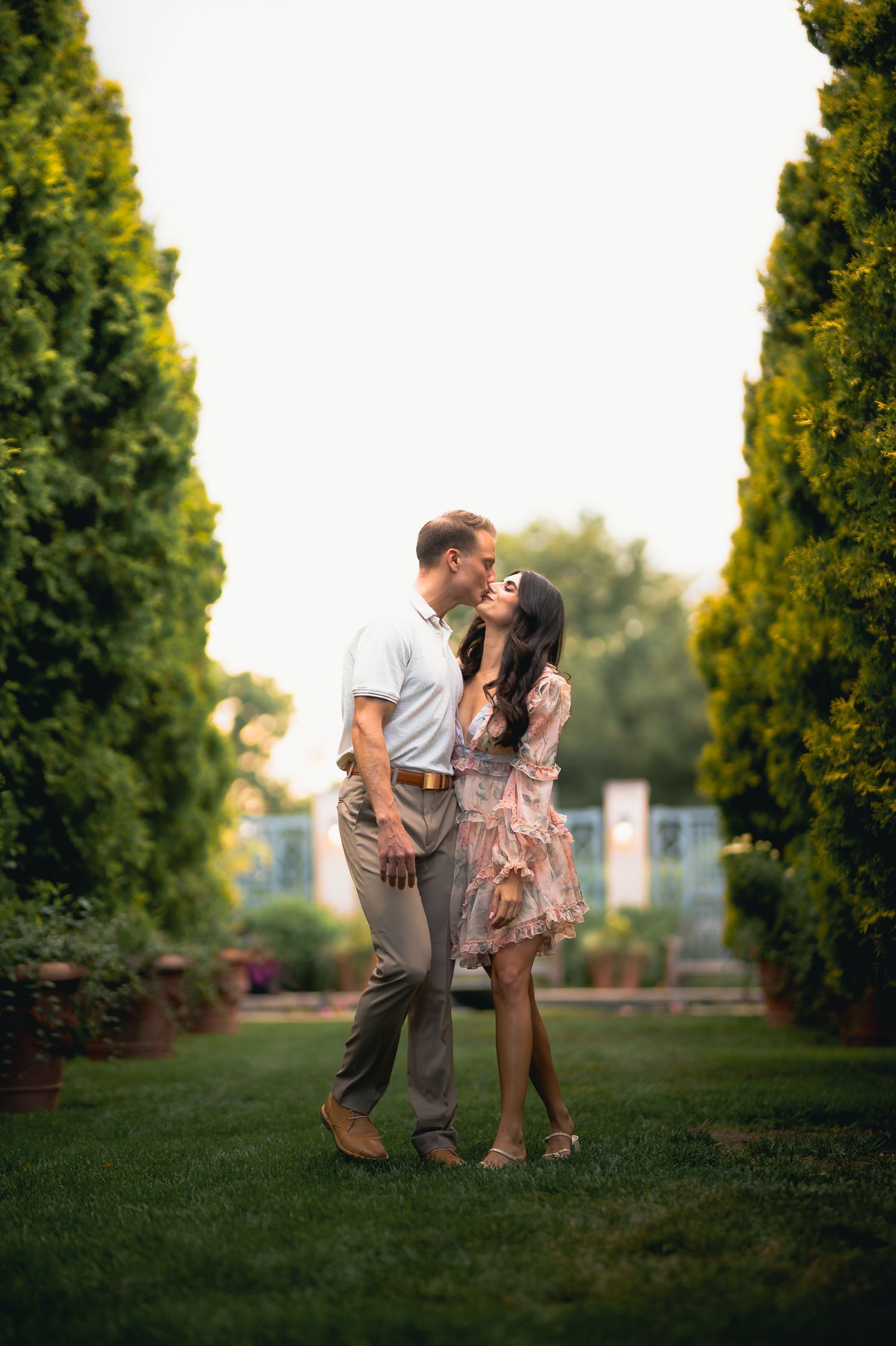 Couple sharing romantic kiss while standing between tall cypress trees in garden.