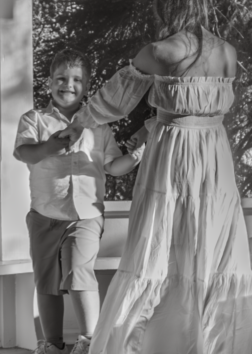 A smiling boy in a button-up shirt dances with a woman in a flowing white dress outdoors in black and white.