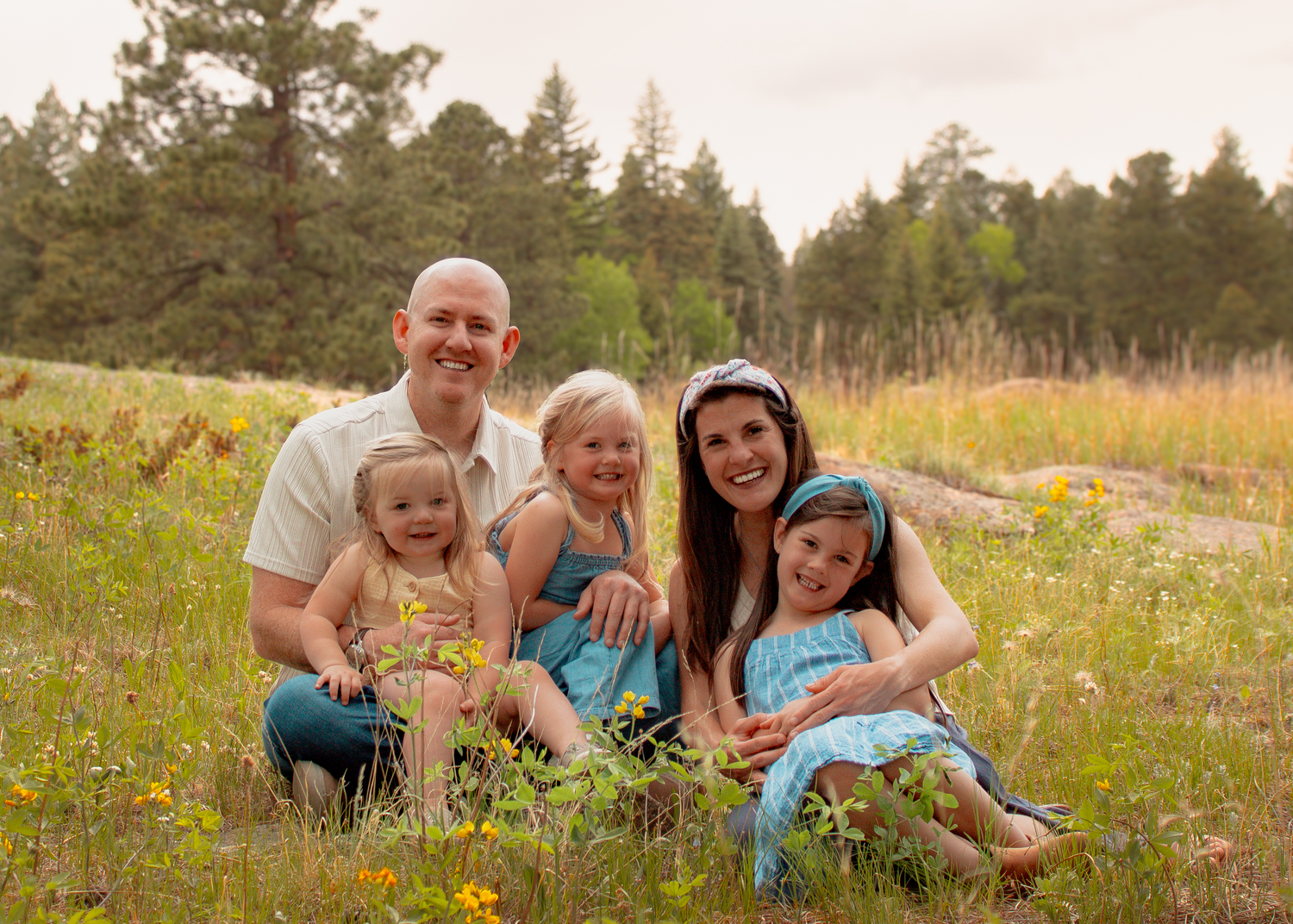Happy family of five smiling together in a sunny wildflower meadow with pine trees in the background.