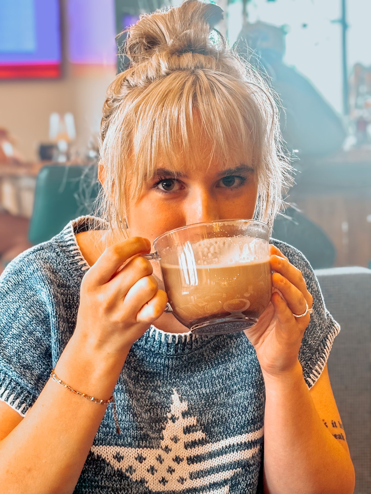 Blonde woman with bangs sipping latte from glass mug in casual cafe setting.