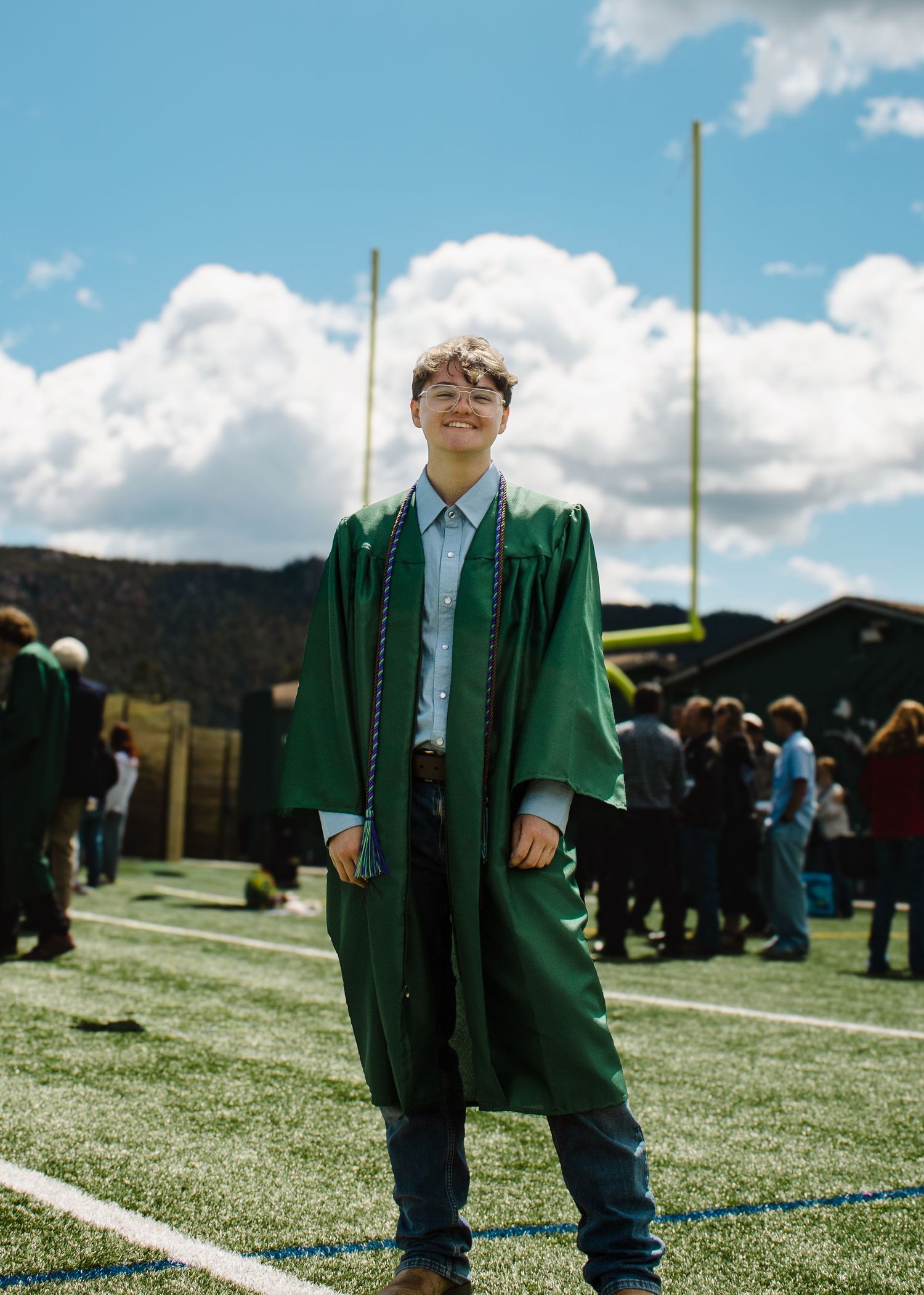 Proud graduate in green gown standing confidently on football field under blue sky at graduation ceremony.