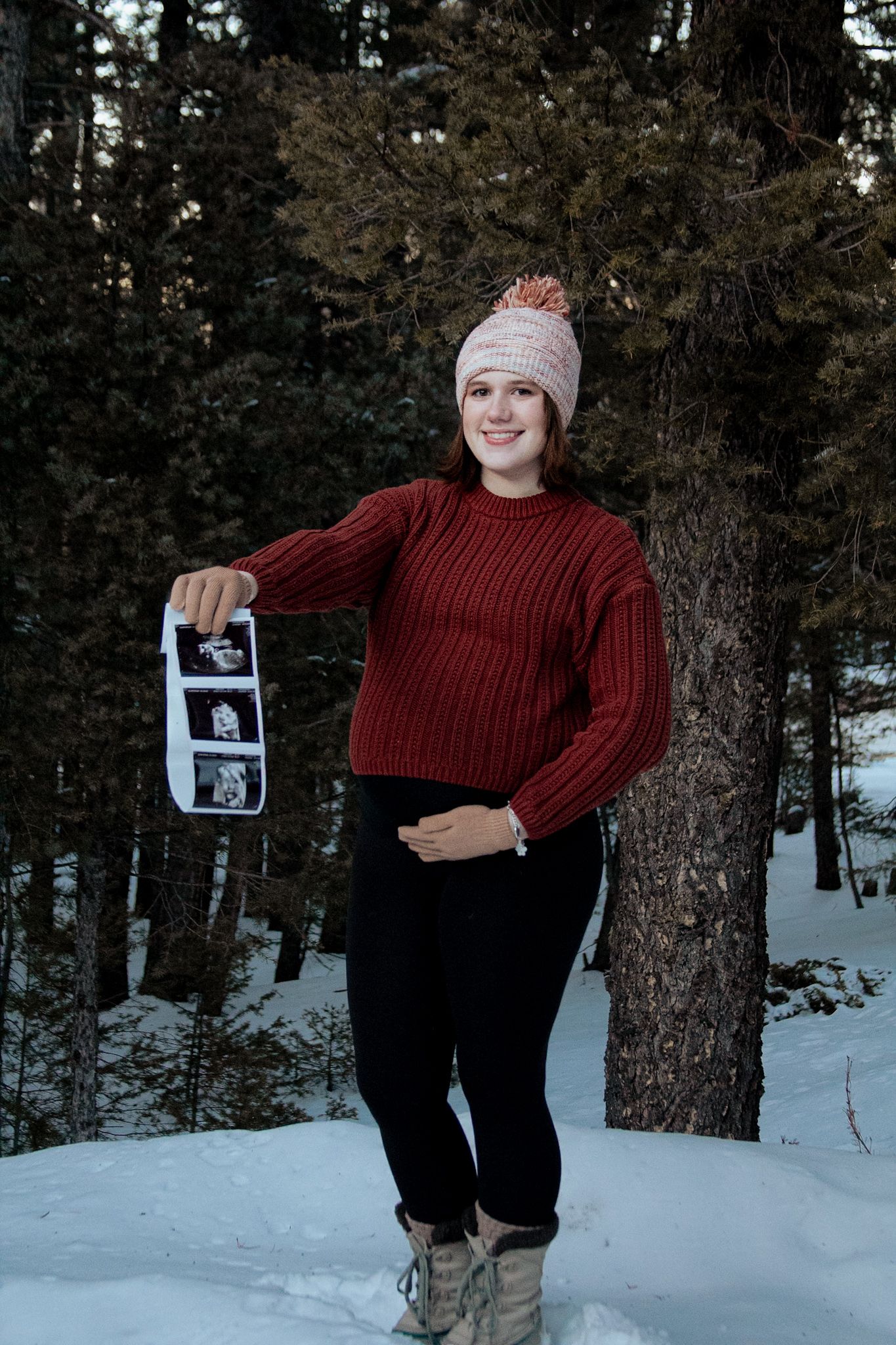Pregnant woman in red sweater and pink beanie holds ultrasound photos outdoors in snowy winter forest setting.