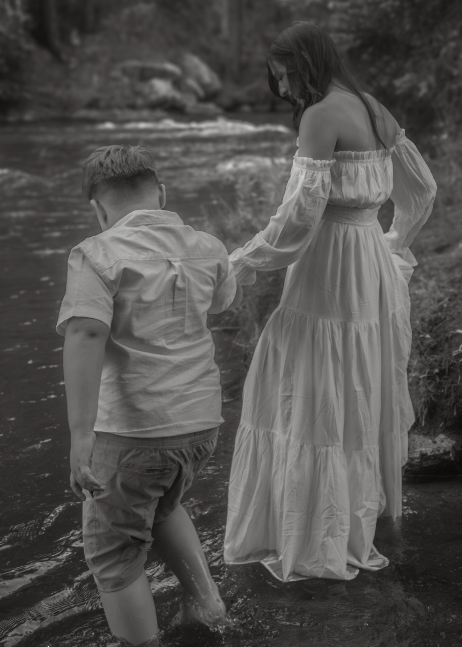 Black and white photo of a woman in a white dress and a young boy wading in a shallow creek together.