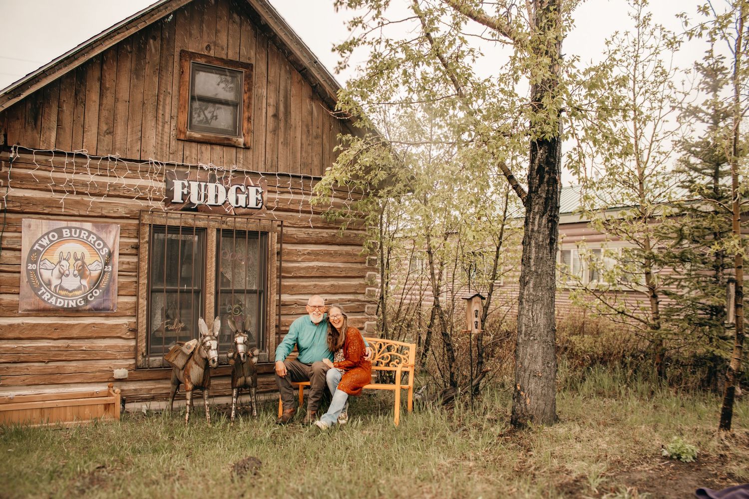 Couple sitting on bench outside rustic log cabin fudge shop with goats nearby.