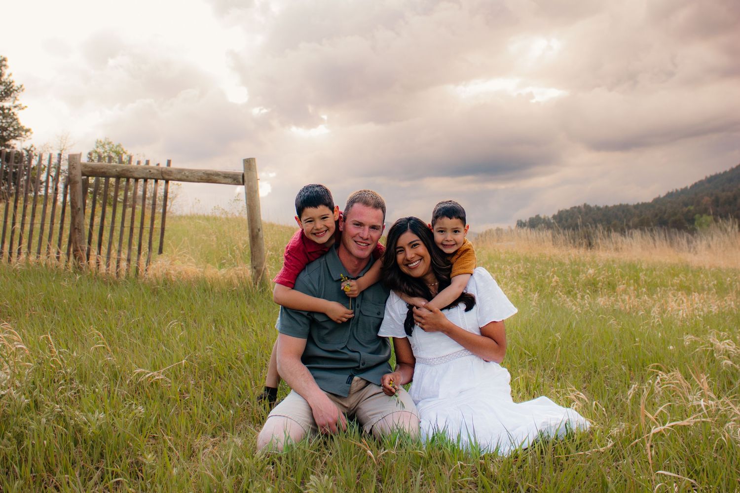 Happy family of four sitting together in a green meadow with dramatic cloudy sky and wooden fence.