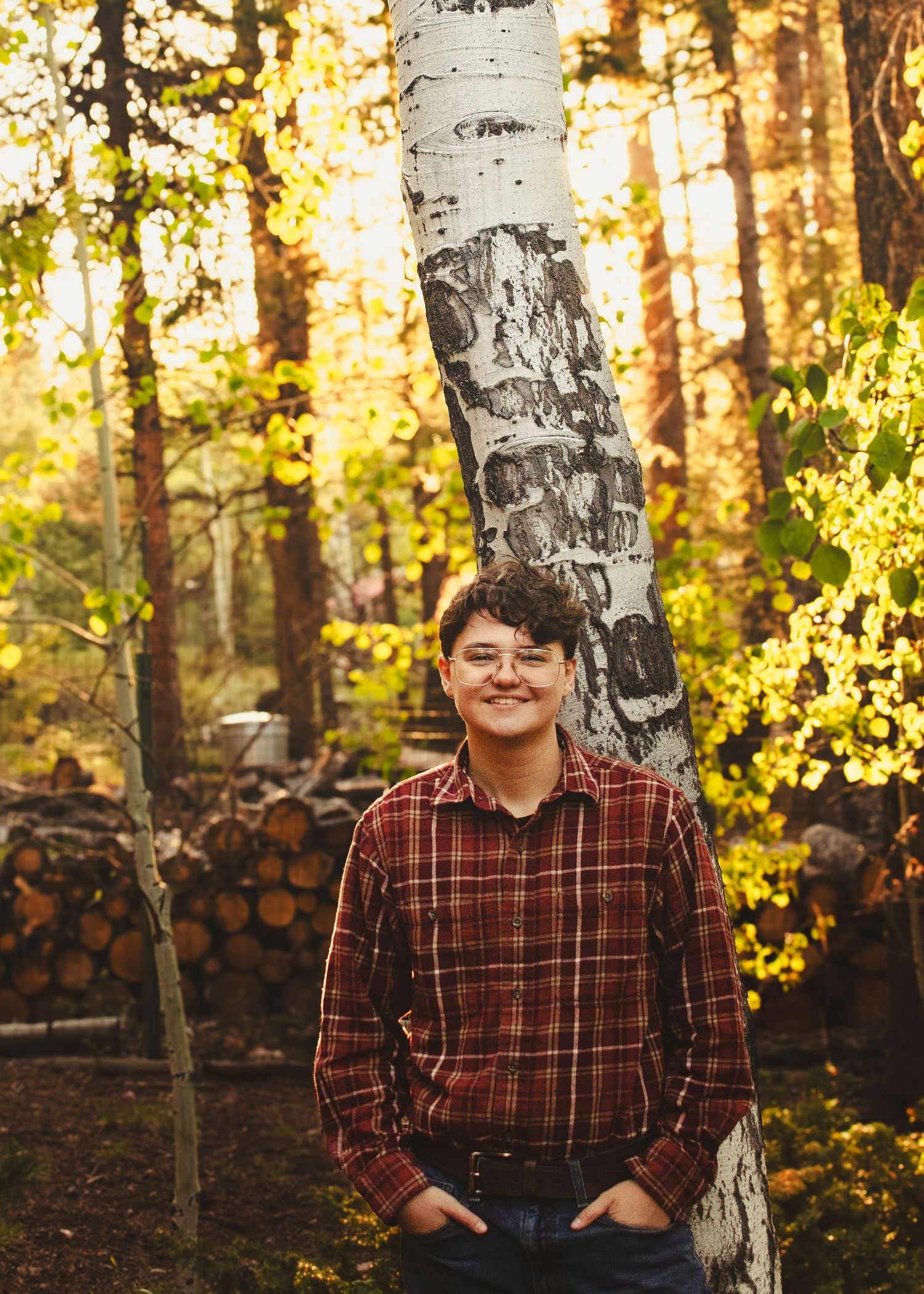 Smiling teen in plaid shirt leaning against birch tree in colorful autumn forest.