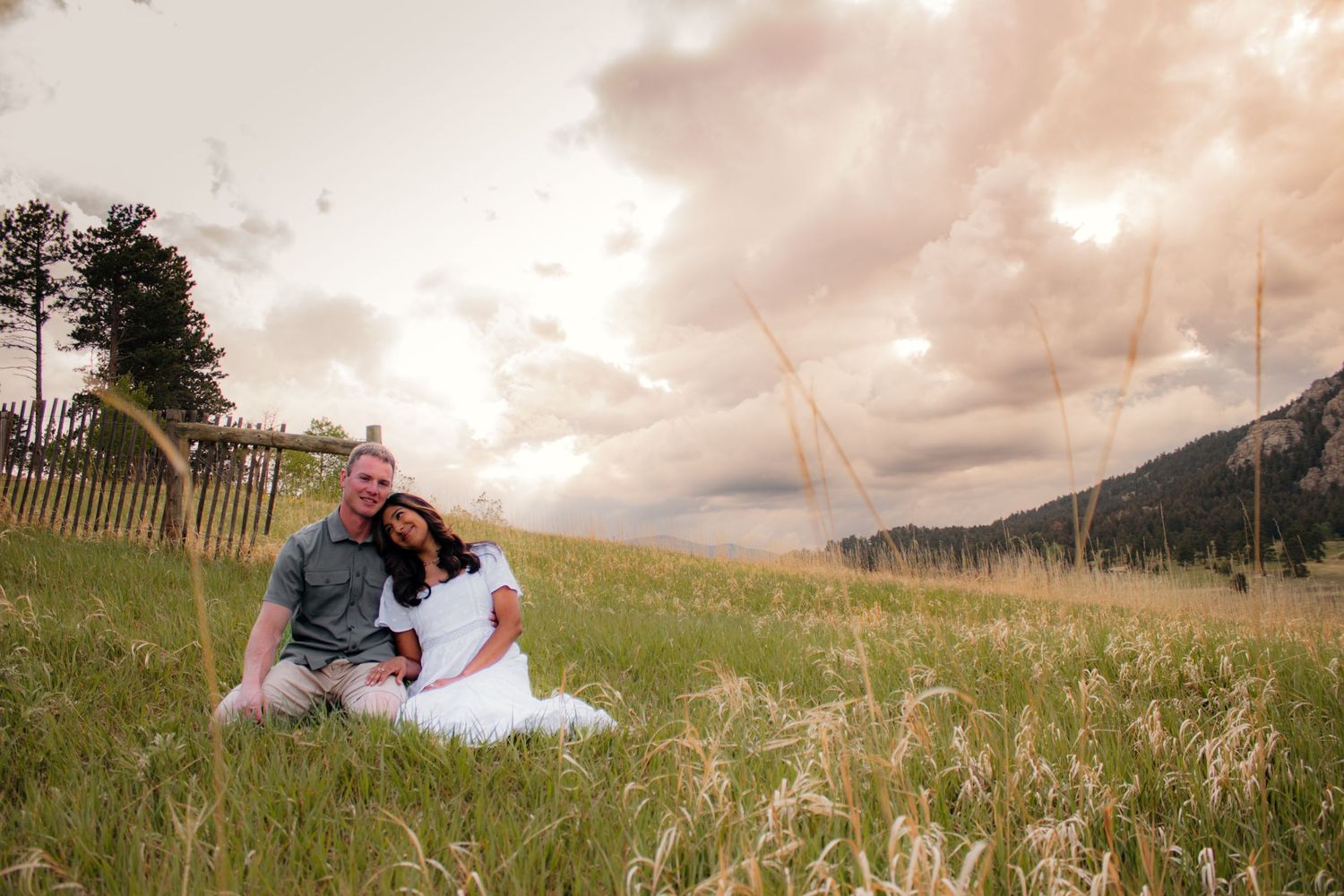 Couple sitting together in a golden grass field with a wooden fence and dramatic cloudy mountain sky.