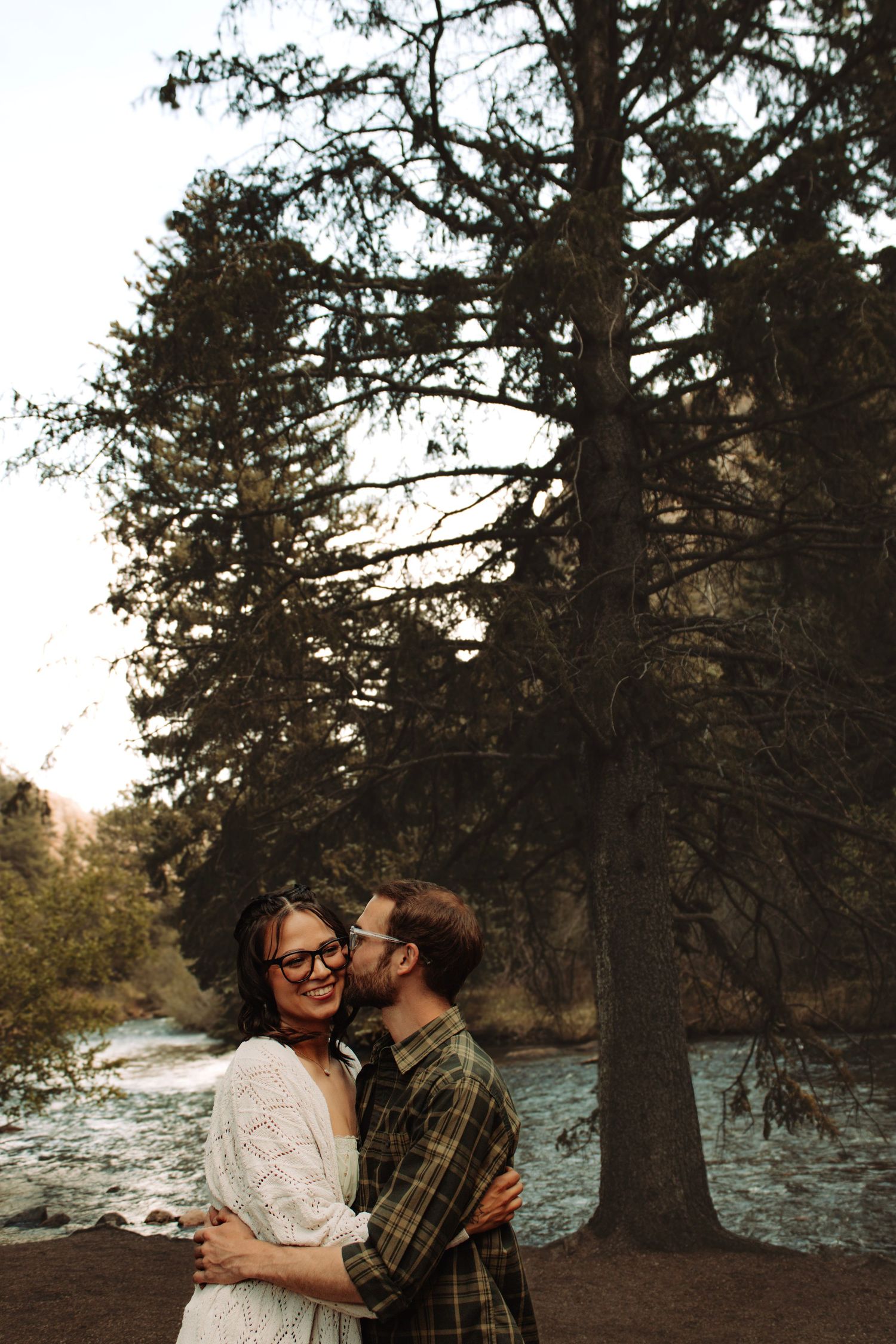 Couple embracing and sharing a kiss near a river surrounded by tall pine trees at golden hour.
