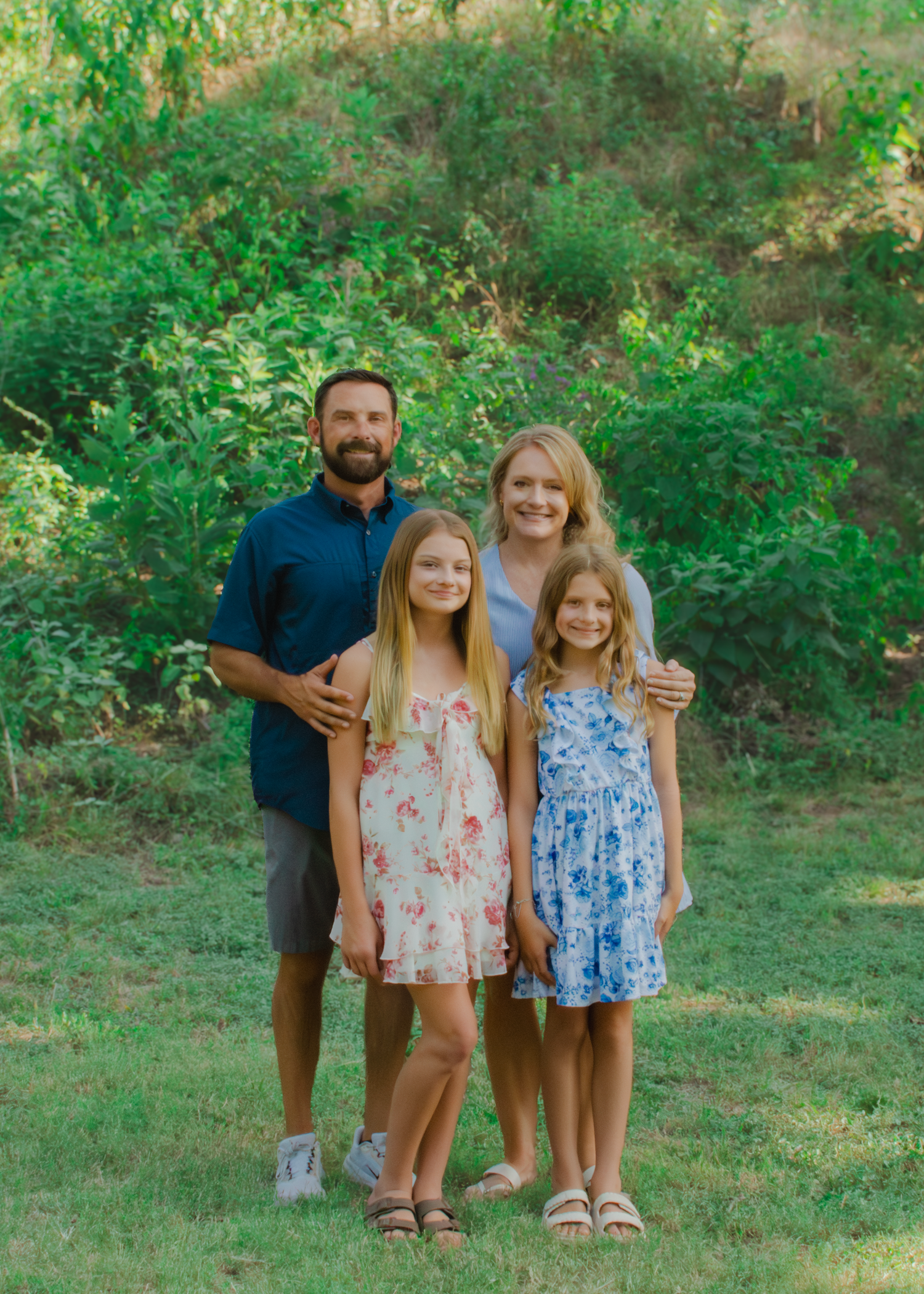 A smiling family of four poses outdoors on green grass with lush trees in the background on a sunny day.