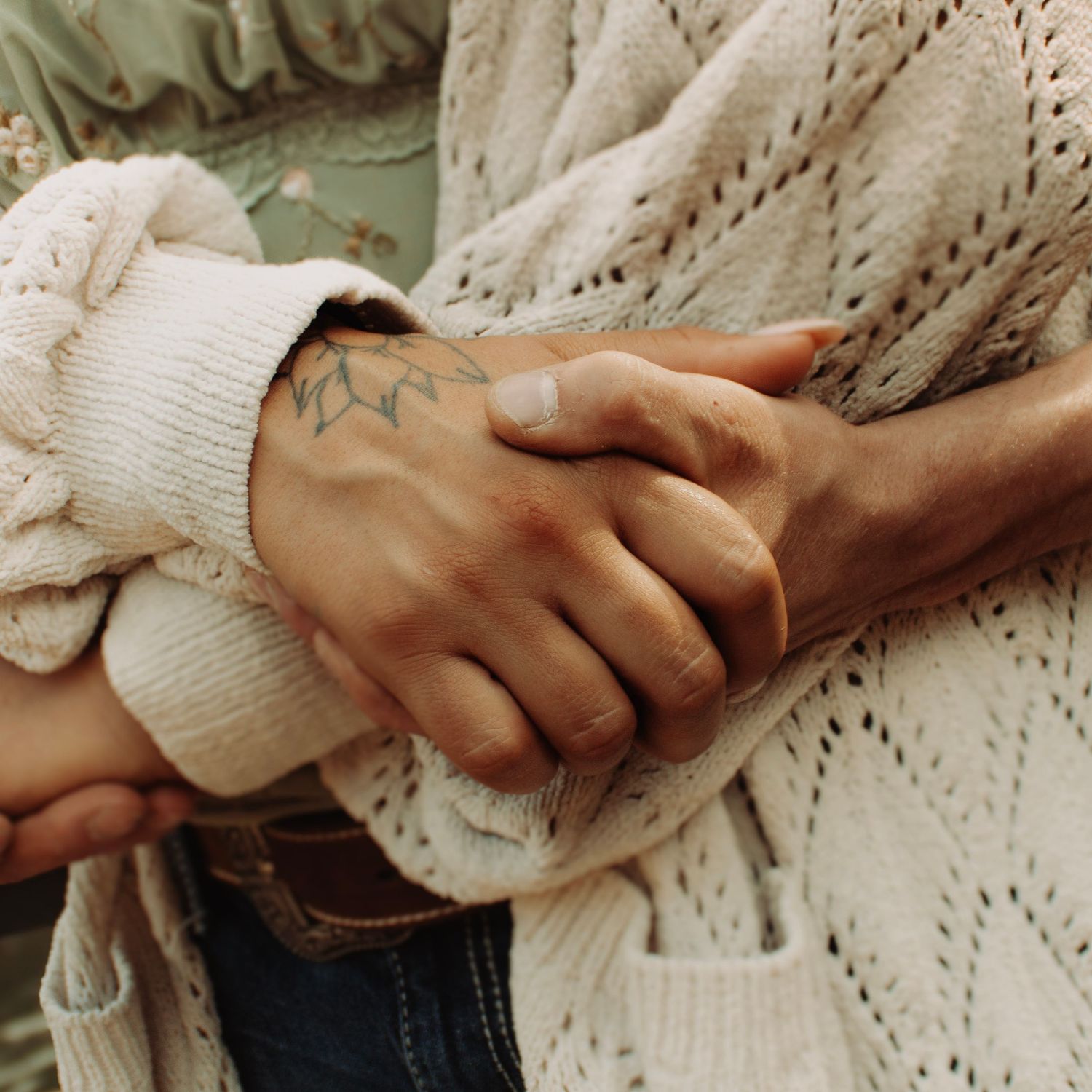 Close-up of two people holding hands, one with a delicate leaf tattoo on their wrist, wearing cozy knit sweaters.