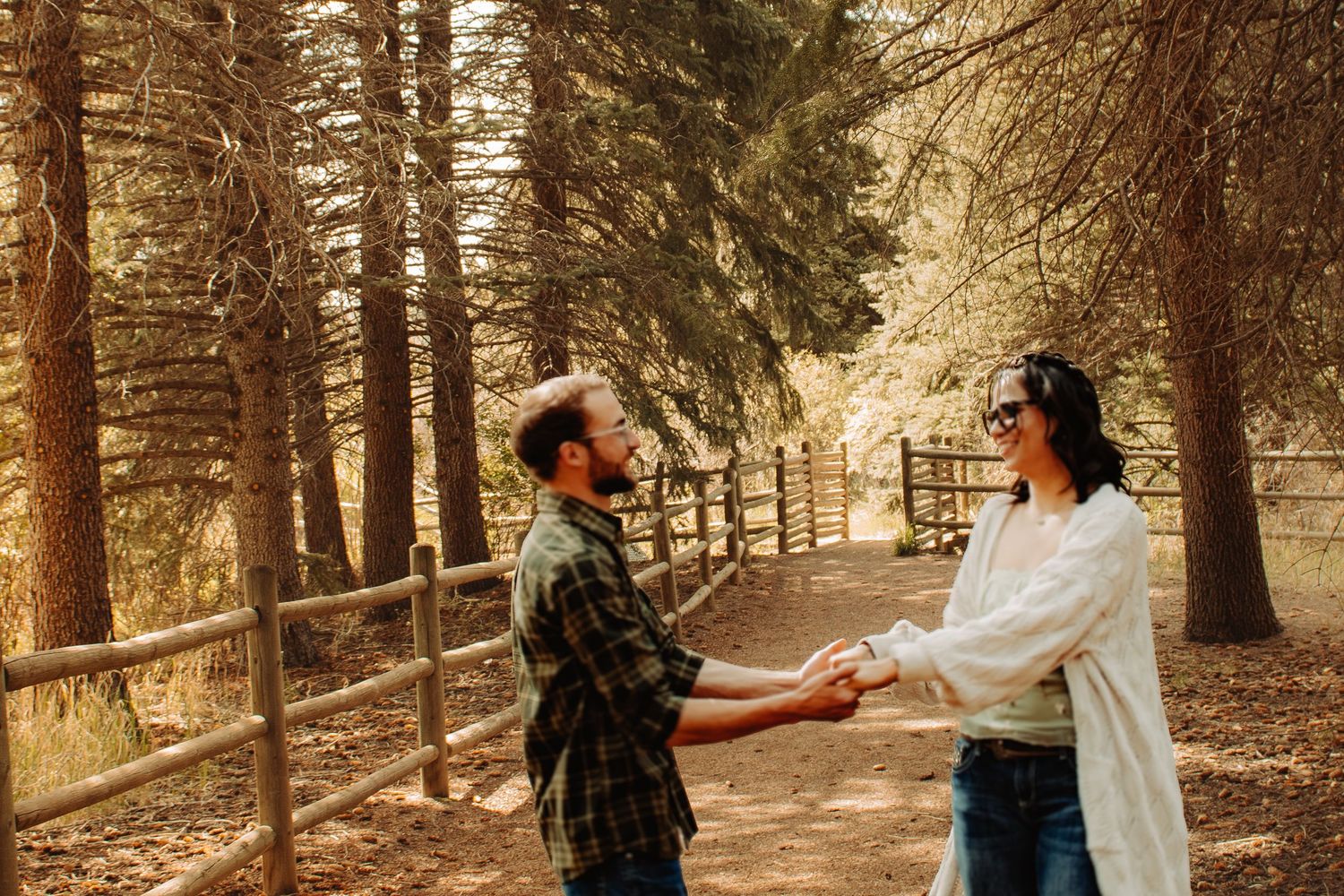 Couple holding hands and facing each other on a forest path lined with pine trees and a wooden fence.