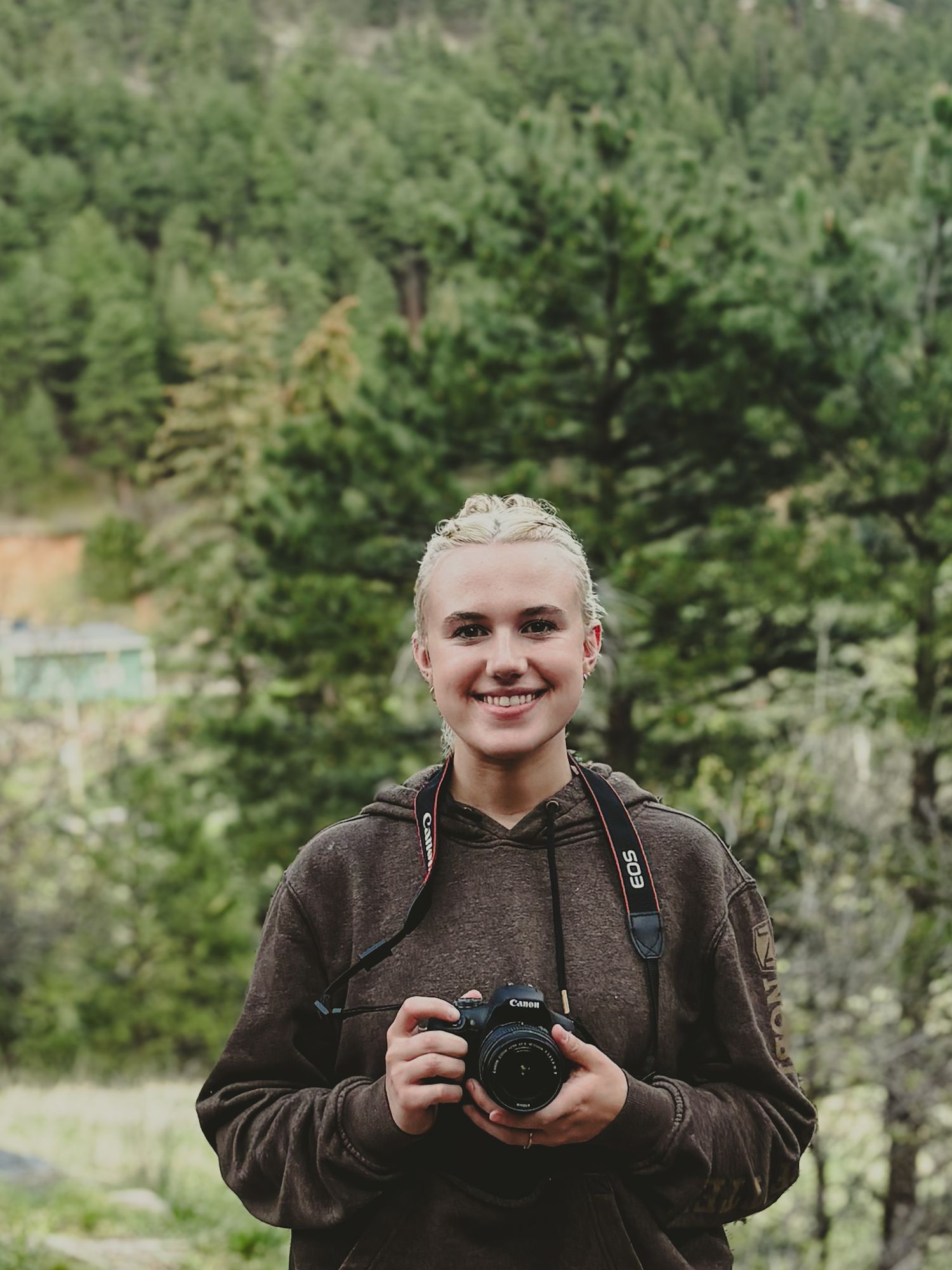 Smiling young photographer holding Canon camera in forest wearing brown hoodie.