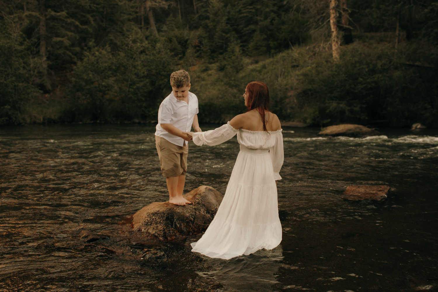 Couple in white attire holding hands in a rocky river surrounded by lush green forest.