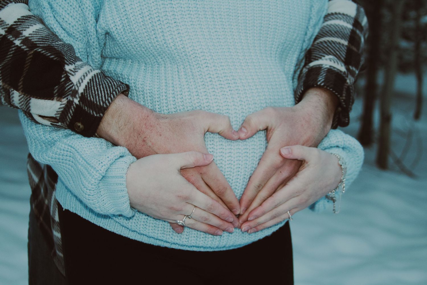 Couple forming a heart shape with hands over pregnant belly outdoors in winter snow.