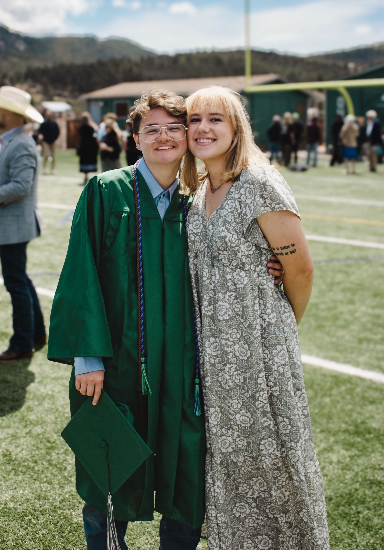 Graduate in green cap and gown smiling with a friend in floral dress at outdoor graduation ceremony.