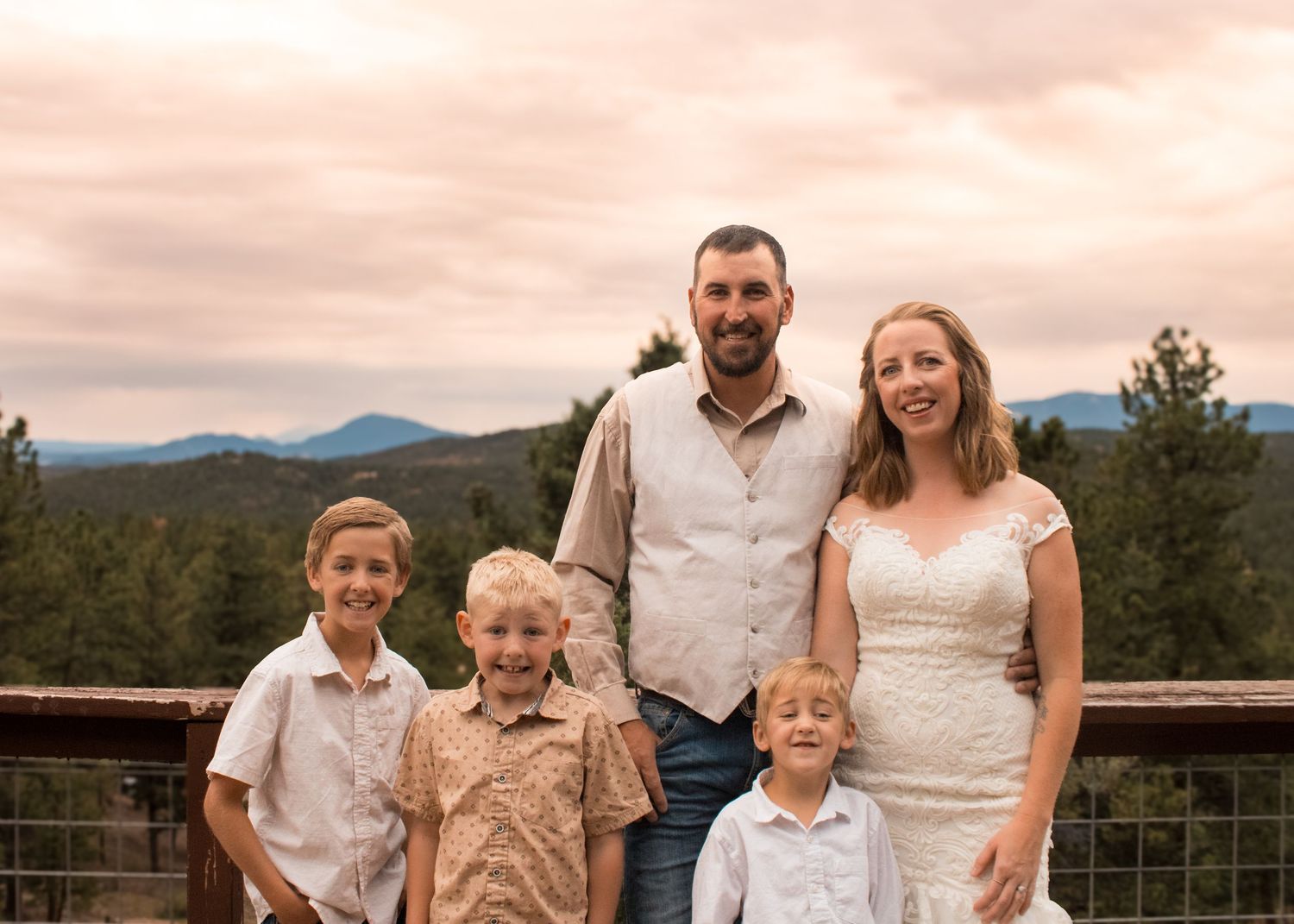 Family of five posing outdoors on a deck with mountain and forest views at sunset.