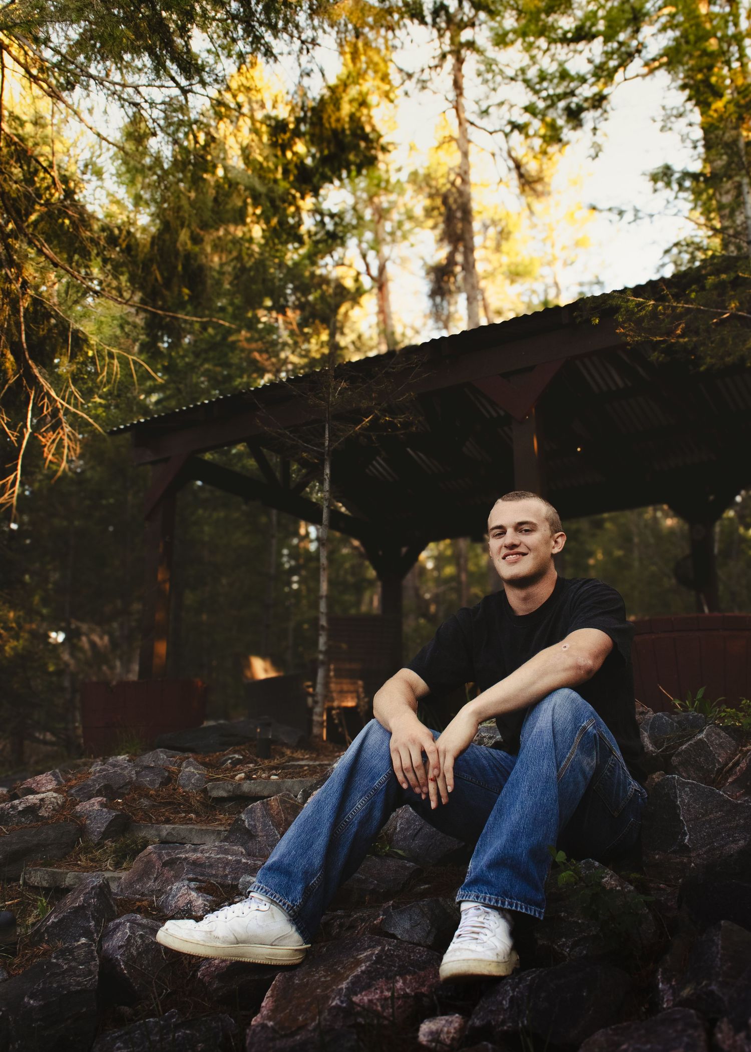 A smiling young man in a black shirt and jeans sits relaxed on rocks near a wooden shelter in a forest.