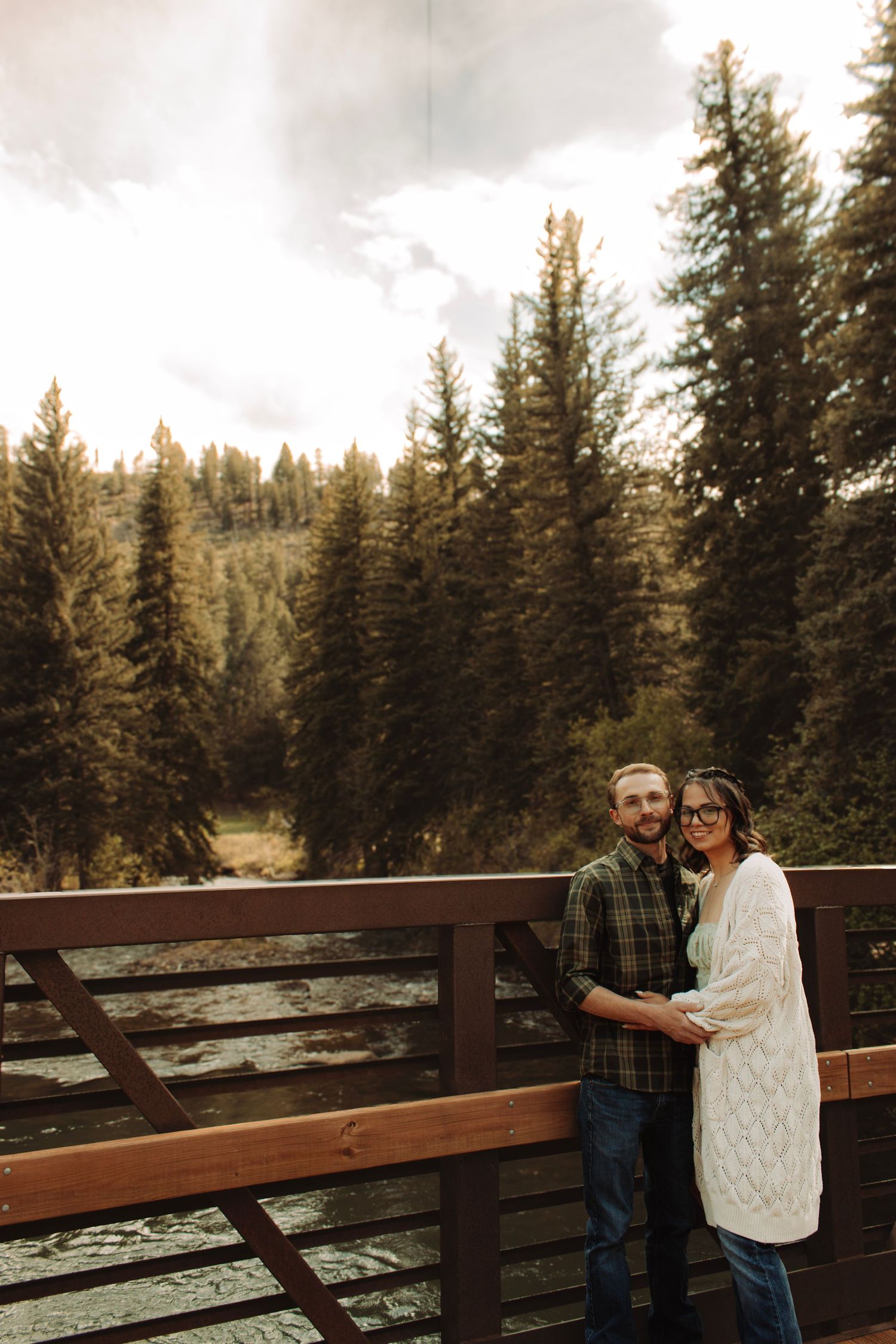 Loving couple posing on a wooden bridge with lush pine forest and mountain stream backdrop.