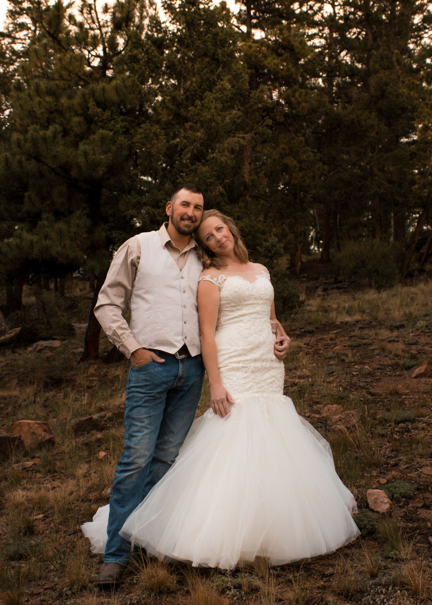 A couple in wedding attire poses outdoors among pine trees, bride in mermaid gown, groom in jeans and vest.