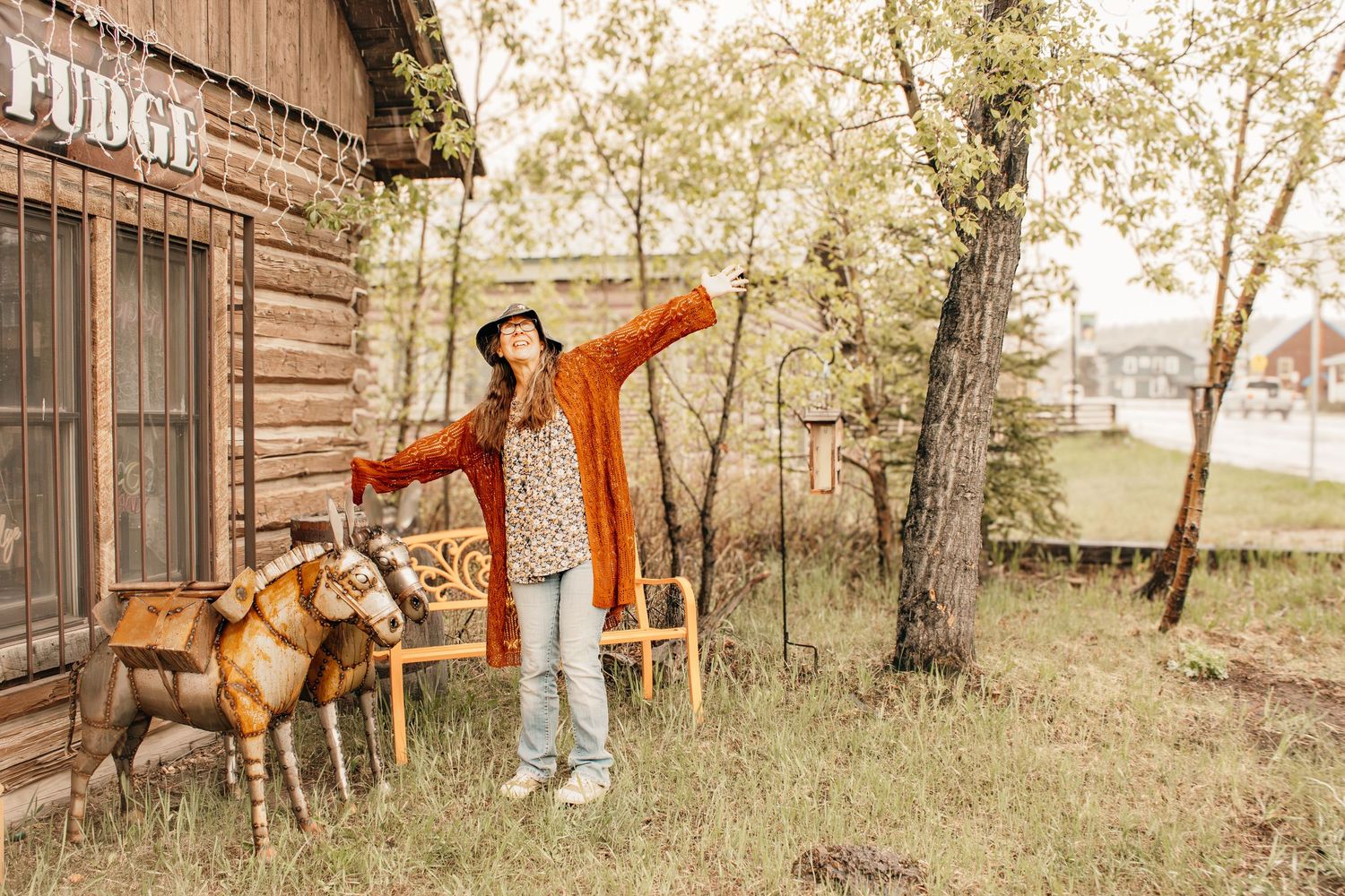Woman in orange cardigan and hat poses with wooden goat sculptures outside a rustic log cabin fudge shop.