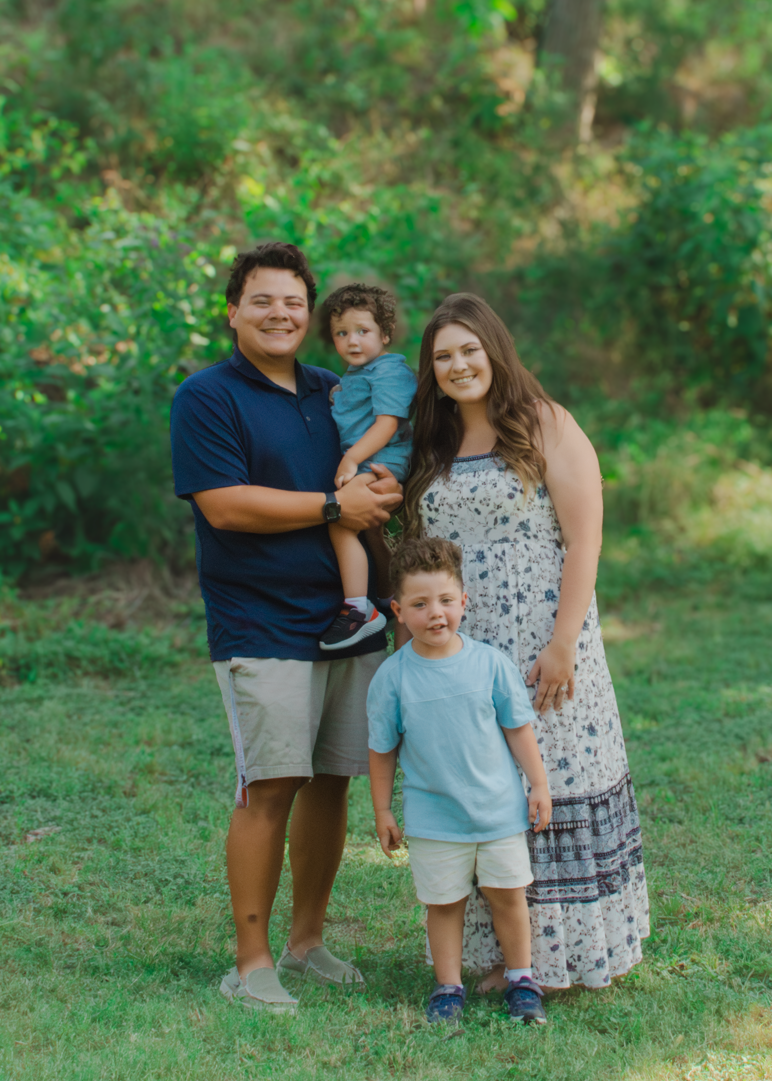 A smiling family of four poses outdoors on green grass, with a man holding a toddler and a woman standing beside a young boy.