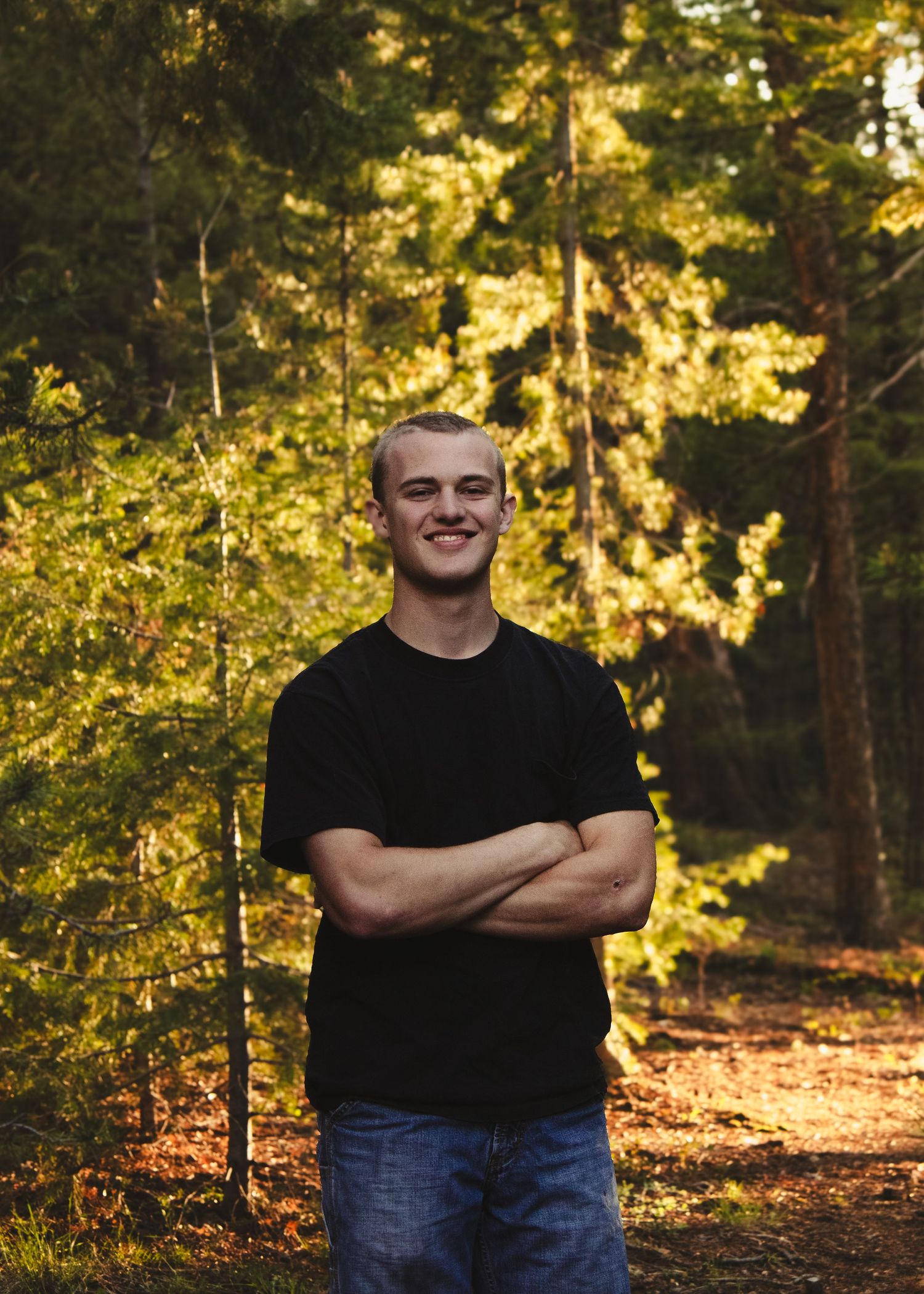 A smiling young man in a black t-shirt stands with arms crossed in a sunlit autumn forest.
