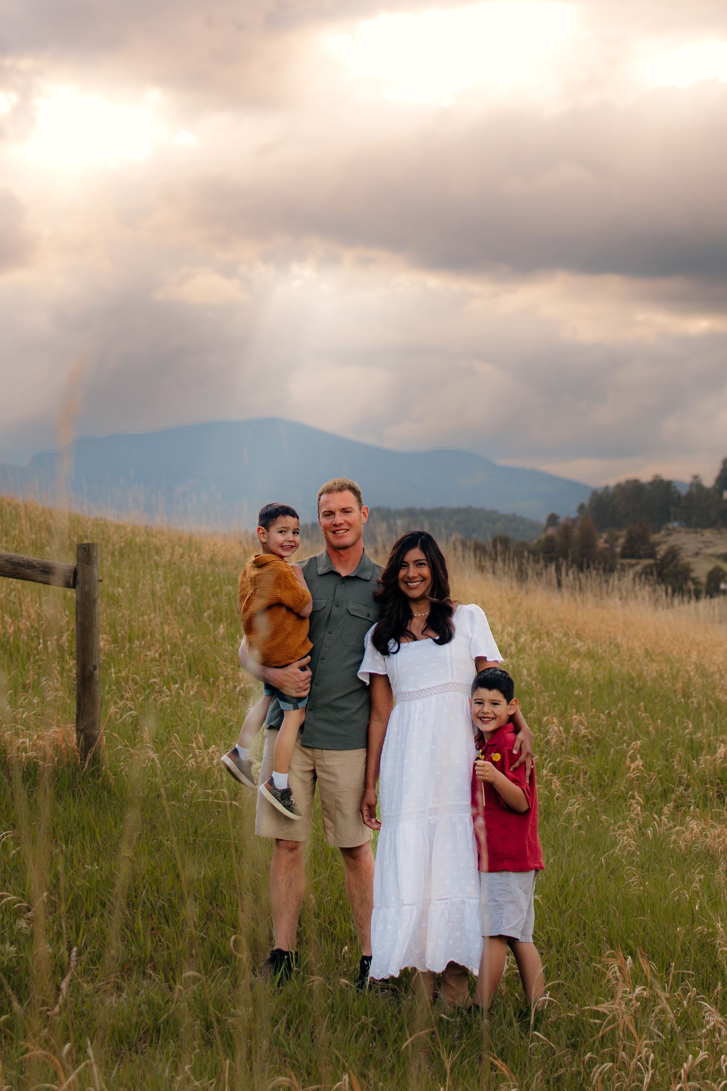 Happy family of four poses in a sunlit mountain meadow with dramatic clouds and golden light behind them.