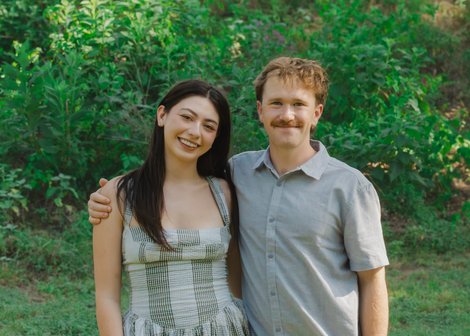 A smiling young couple poses together outdoors in front of lush green trees and foliage.