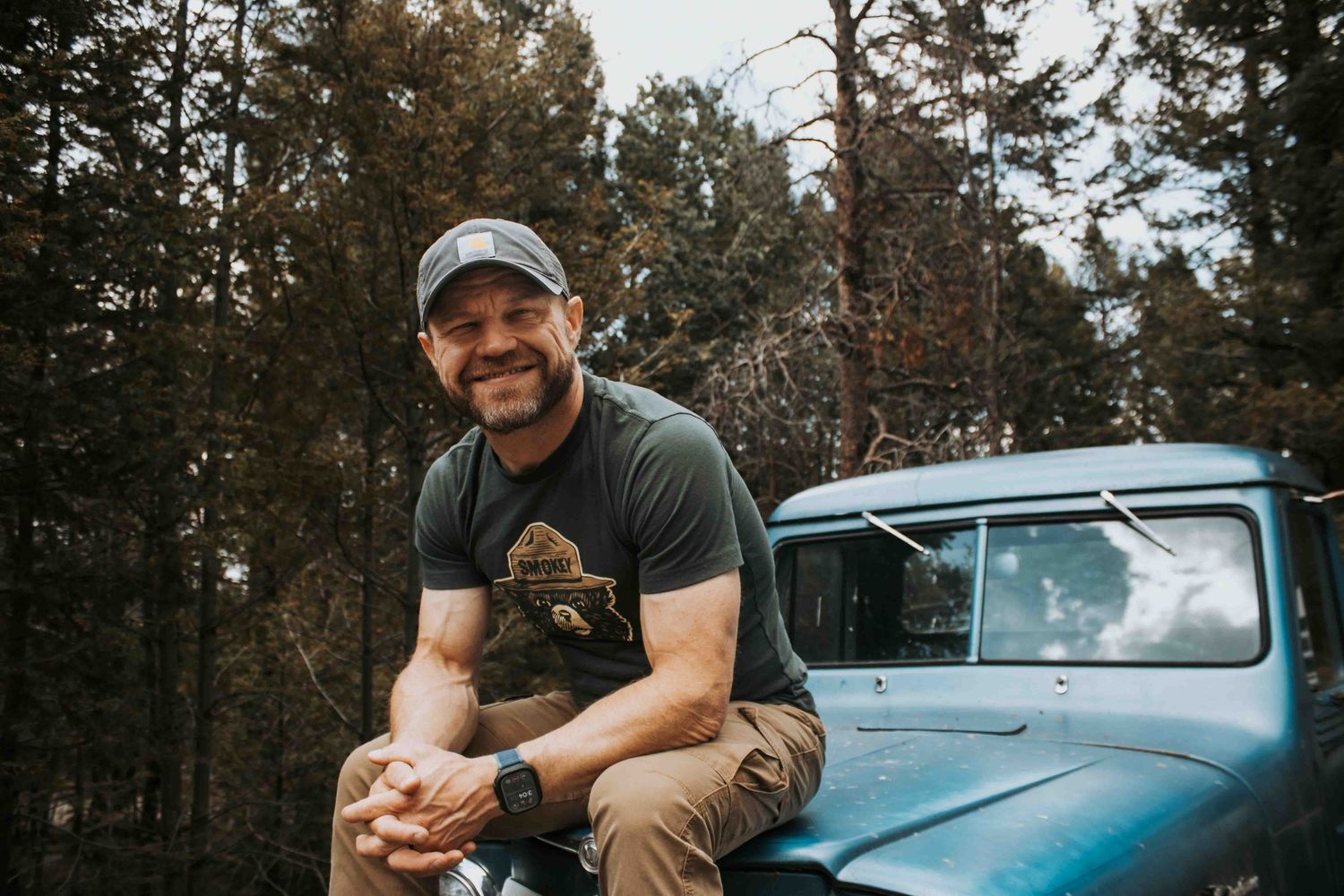 A smiling man in a cap and t-shirt sits on a vintage blue truck surrounded by tall pine trees outdoors.