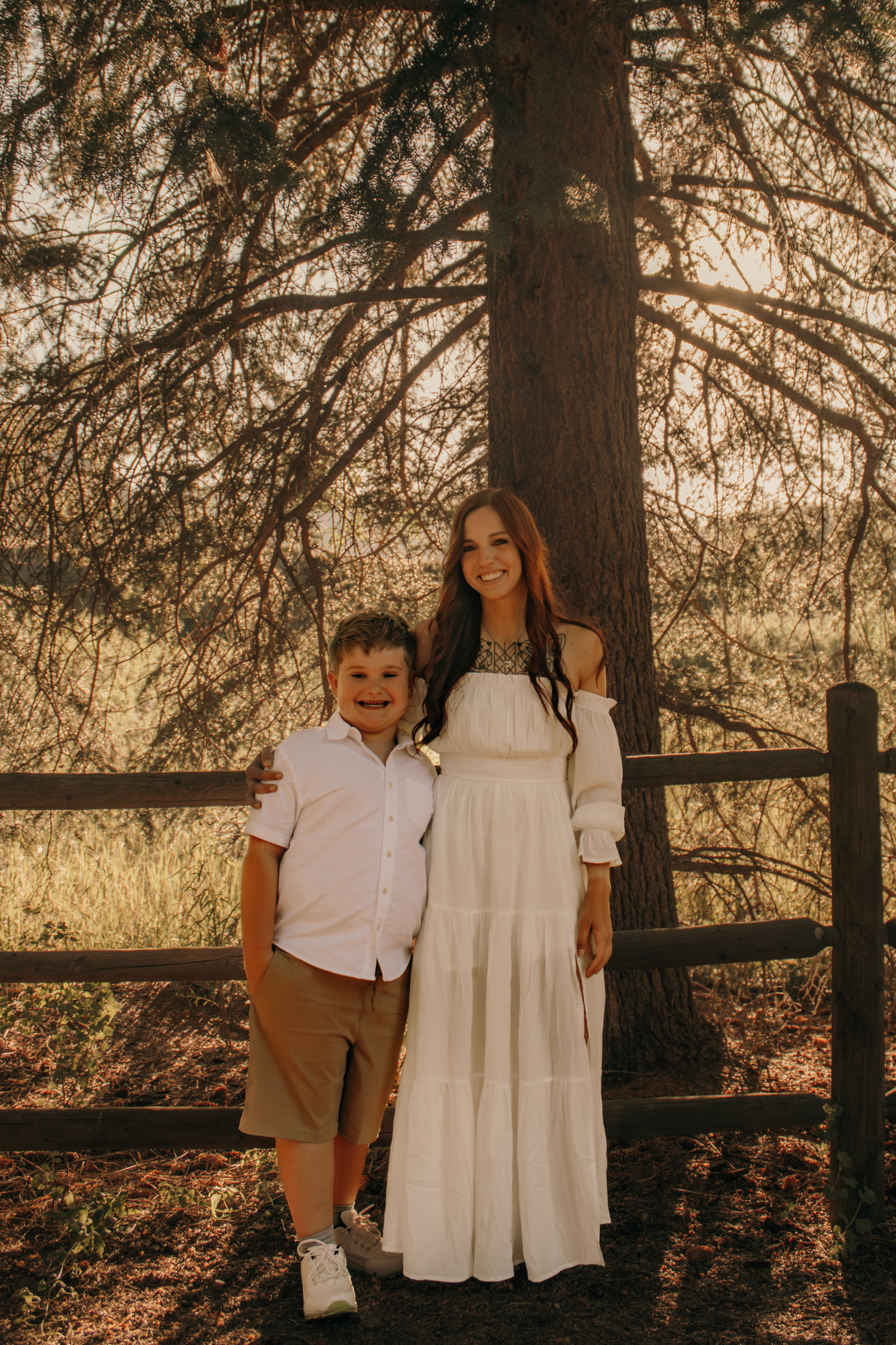 A woman in a white dress and a young boy in white shirt and tan shorts pose by a wooden fence near a pine tree.