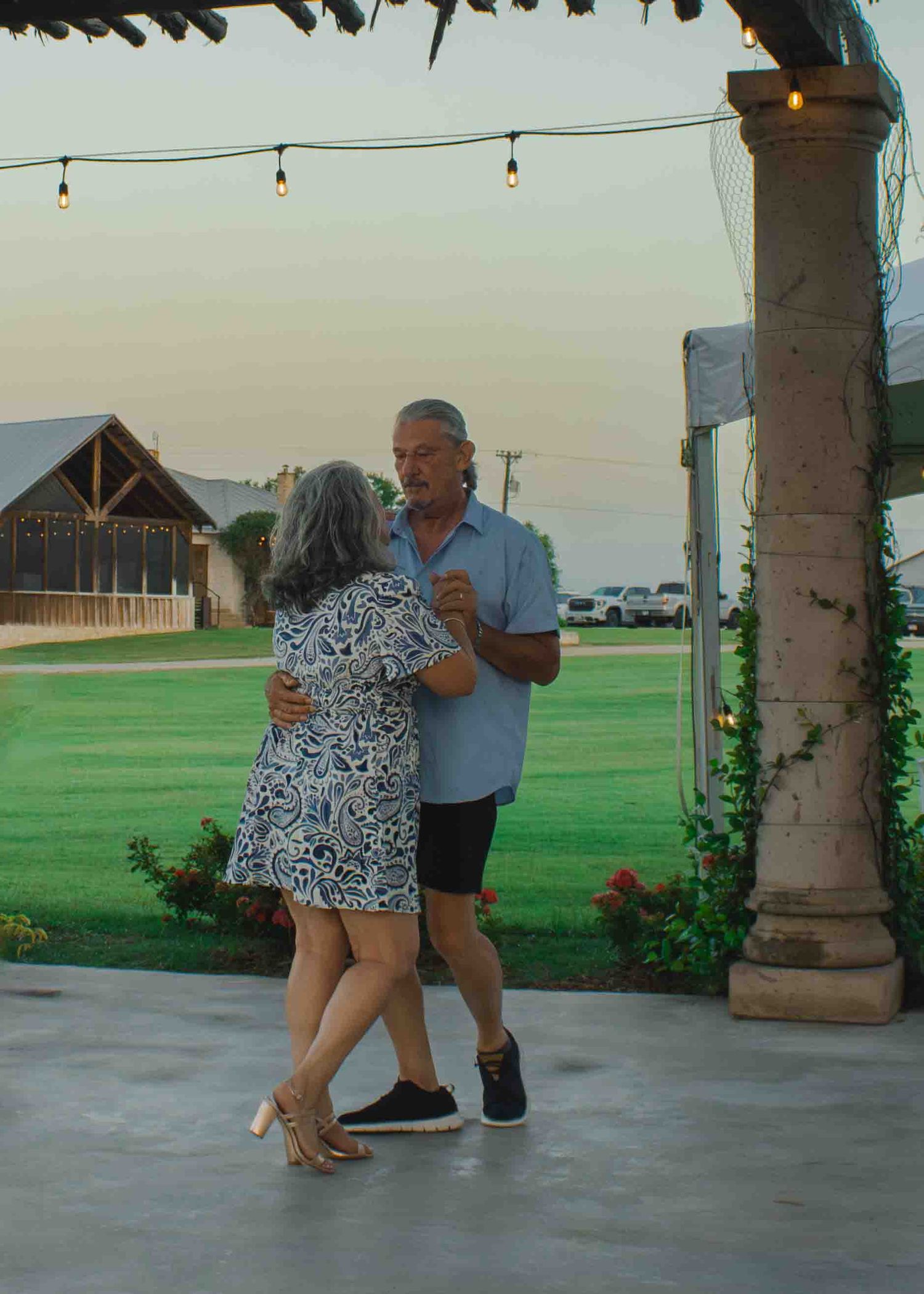Elderly couple dancing outdoors on a patio at dusk, string lights above, green lawn in background.