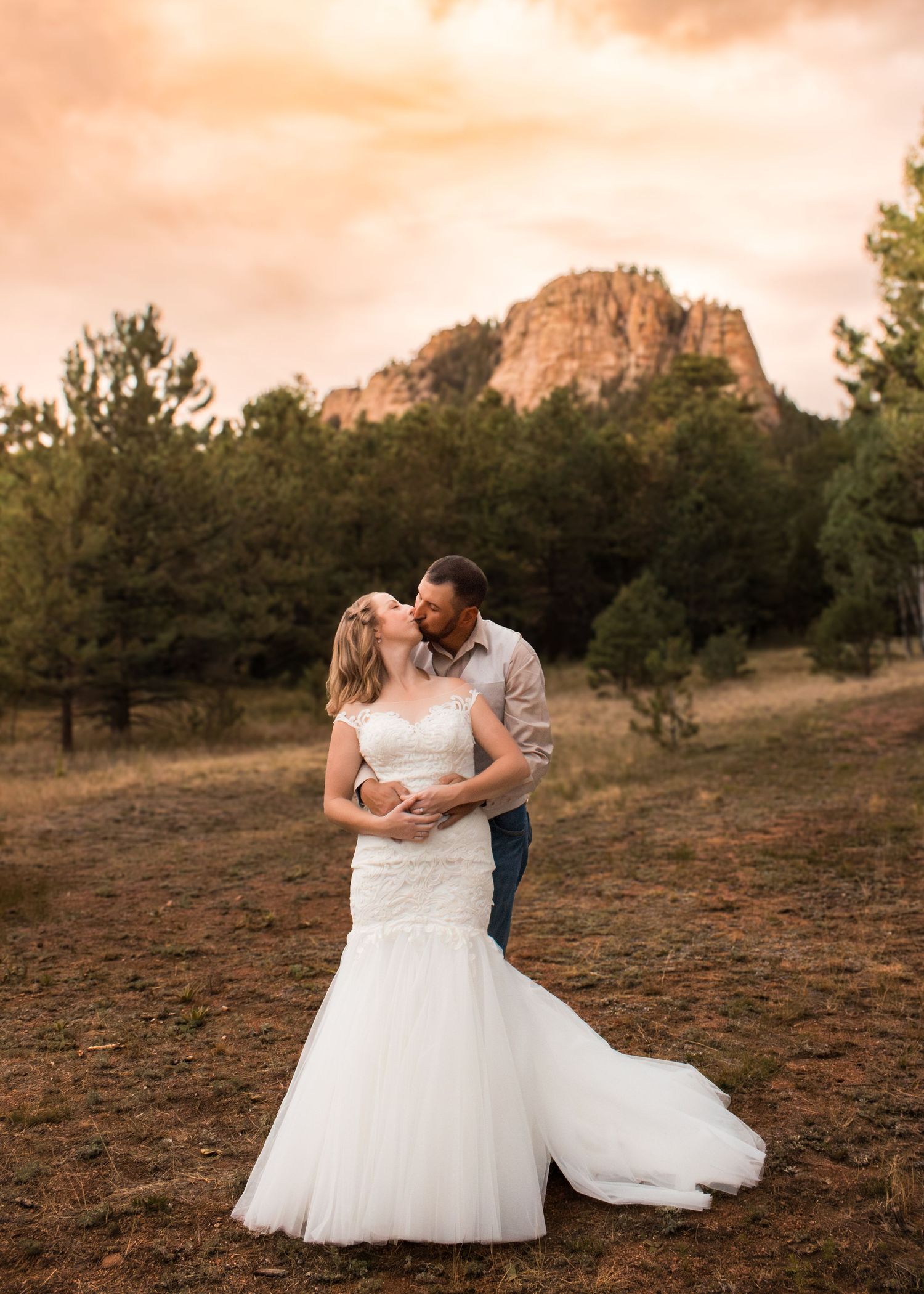Bride and groom share a romantic kiss at sunset with a rocky mountain and pine trees in the background.
