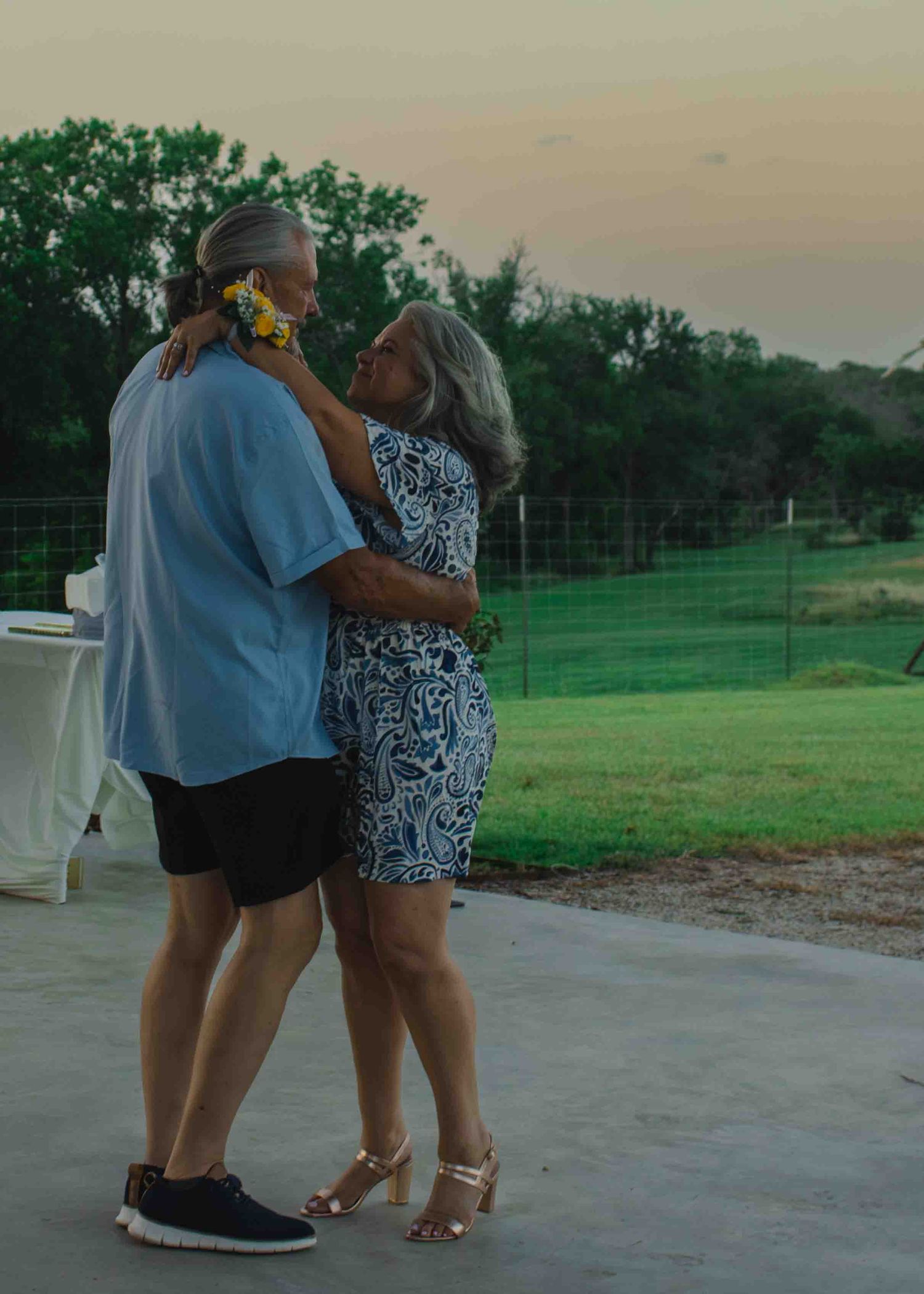 Elderly couple dancing outdoors at sunset near a green fence, woman in floral dress, man in blue shirt.