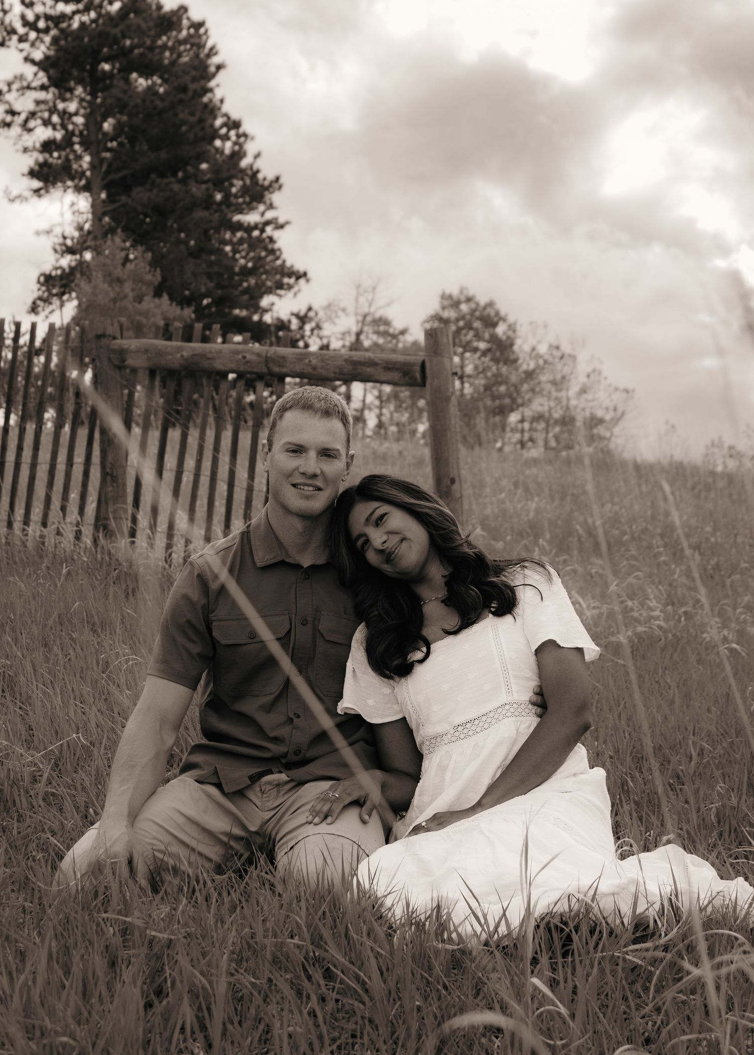 Couple sitting in tall grass near a rustic wooden fence under a cloudy sky in sepia tone.