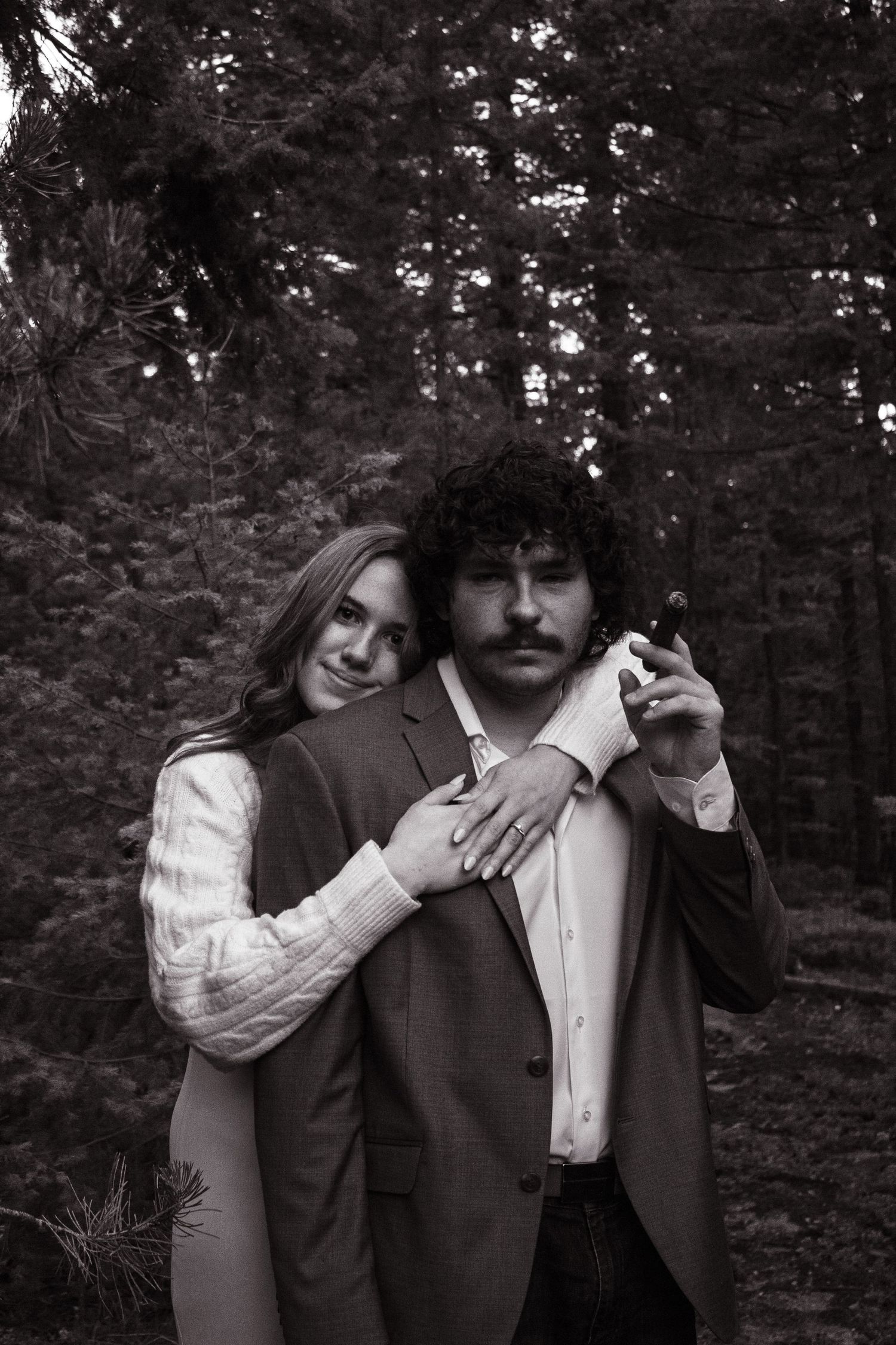 Black and white portrait of a couple embracing in a pine forest, man holding a small object.