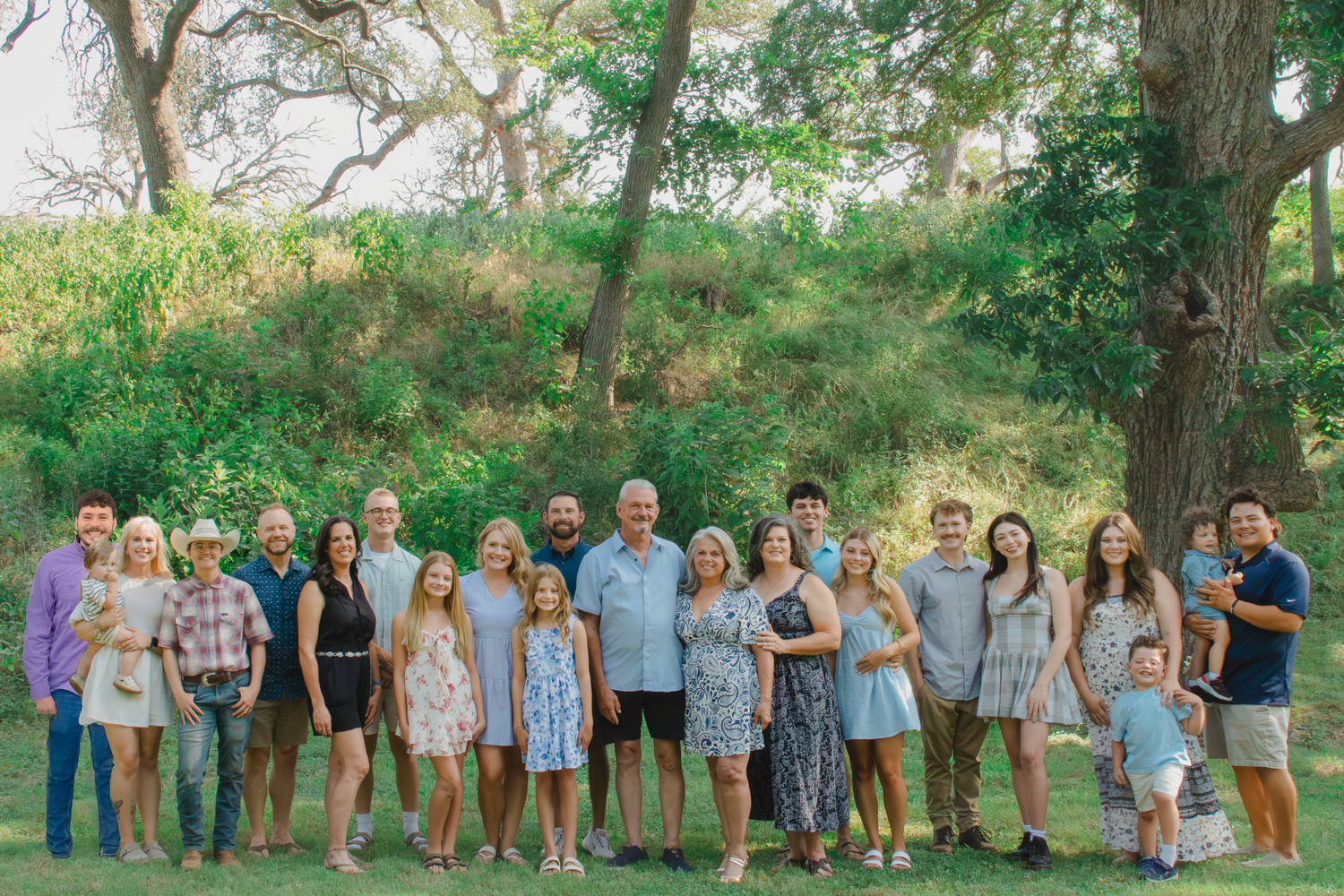 Large family group portrait outdoors, smiling together on green grass with trees in background on a sunny day.