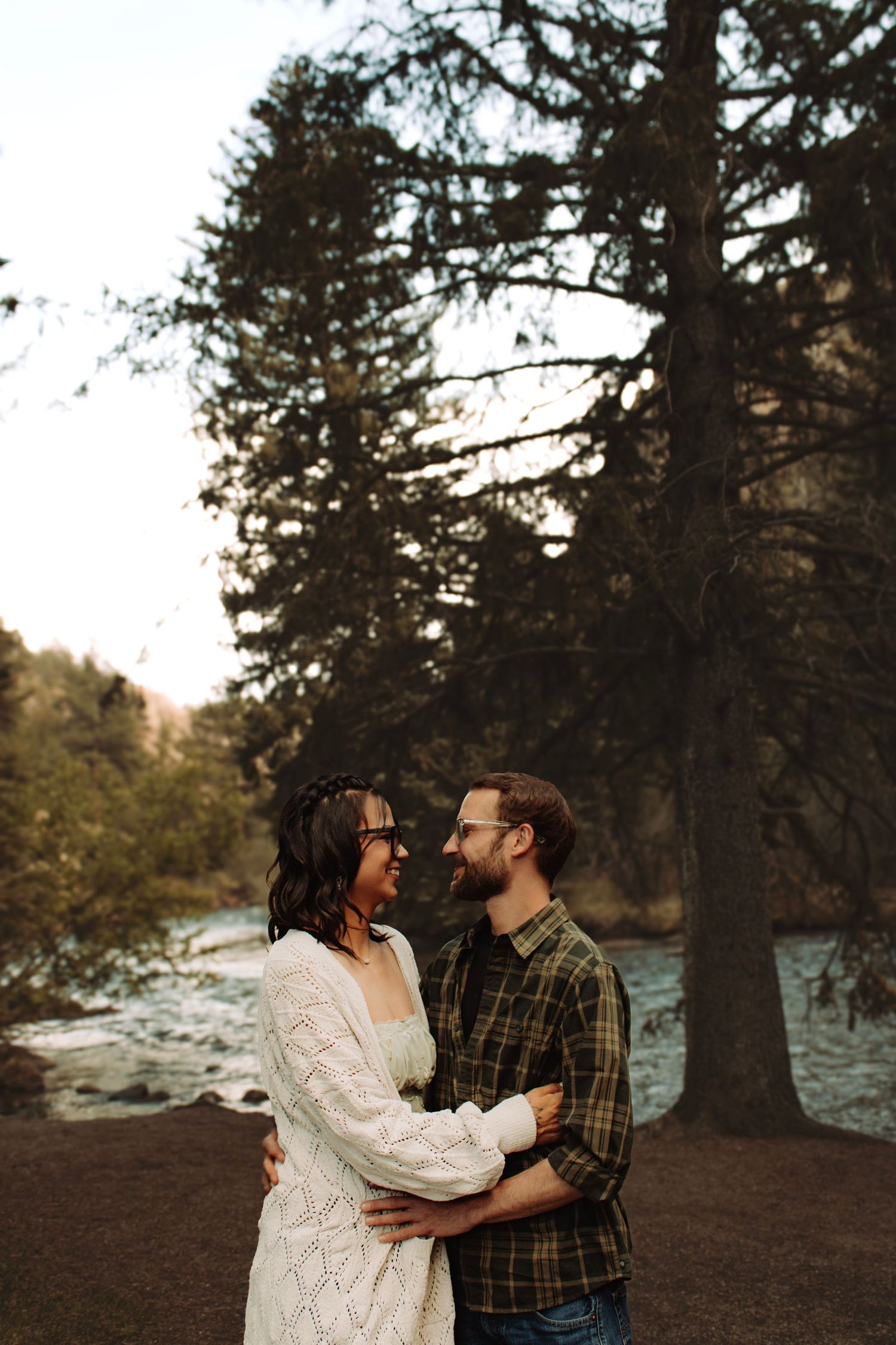 Romantic couple embracing outdoors near a river surrounded by tall evergreen trees at sunset.