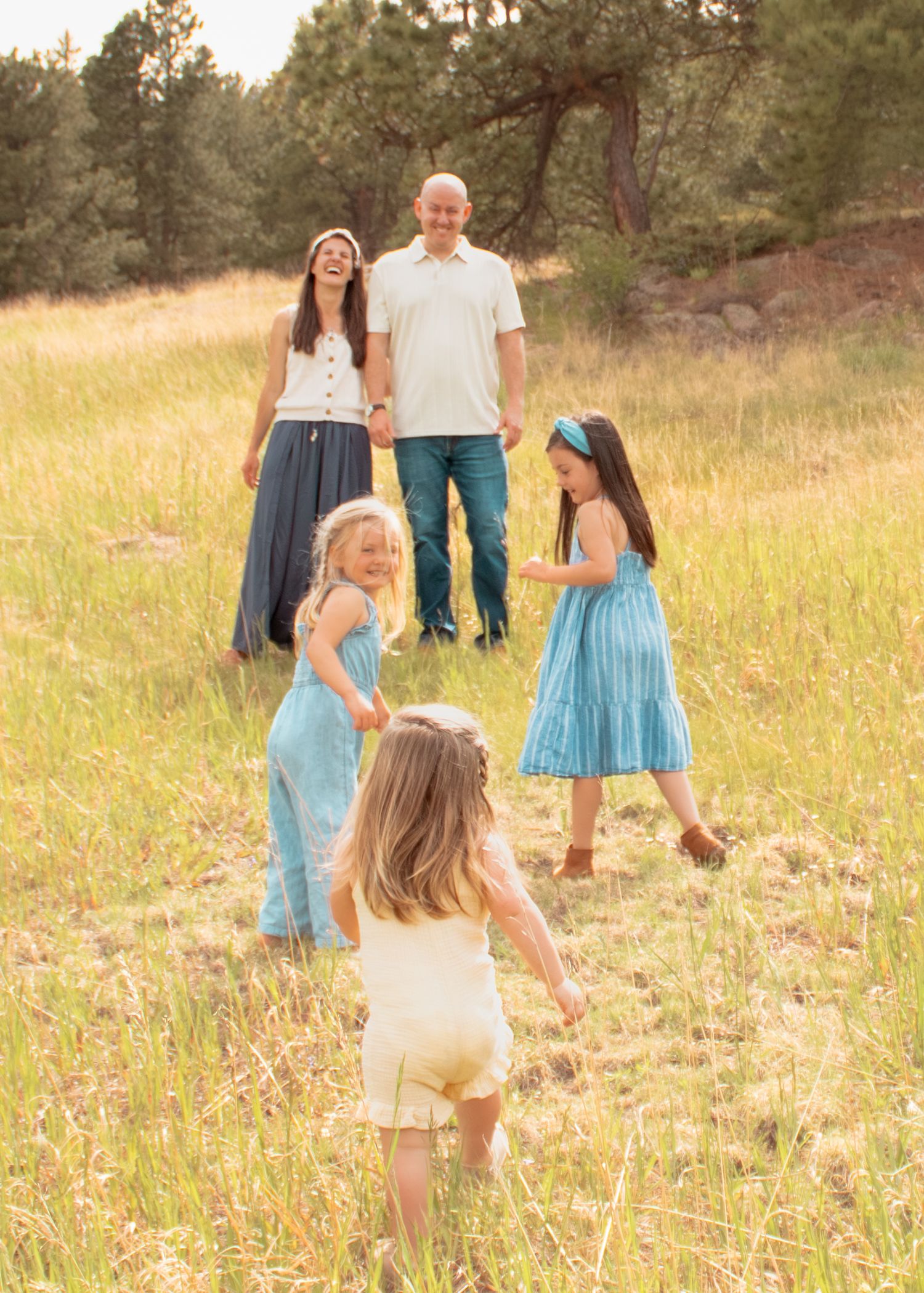 Family of five in a sunny meadow, parents watching three young girls in blue and cream dresses play together outdoors.