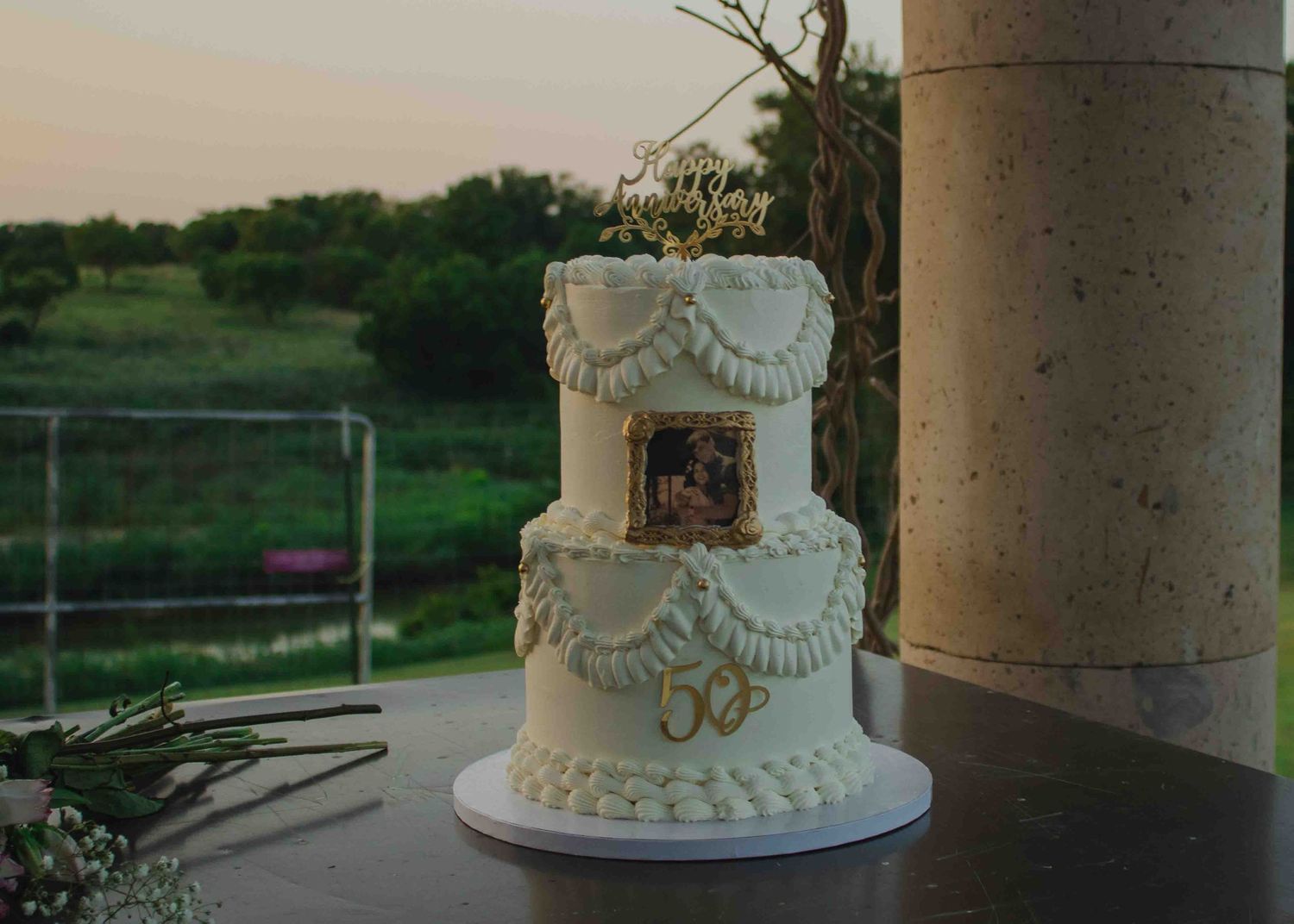 Two-tier white wedding cake with piped ruffles, vintage photo, and gold 50th anniversary topper on outdoor table.