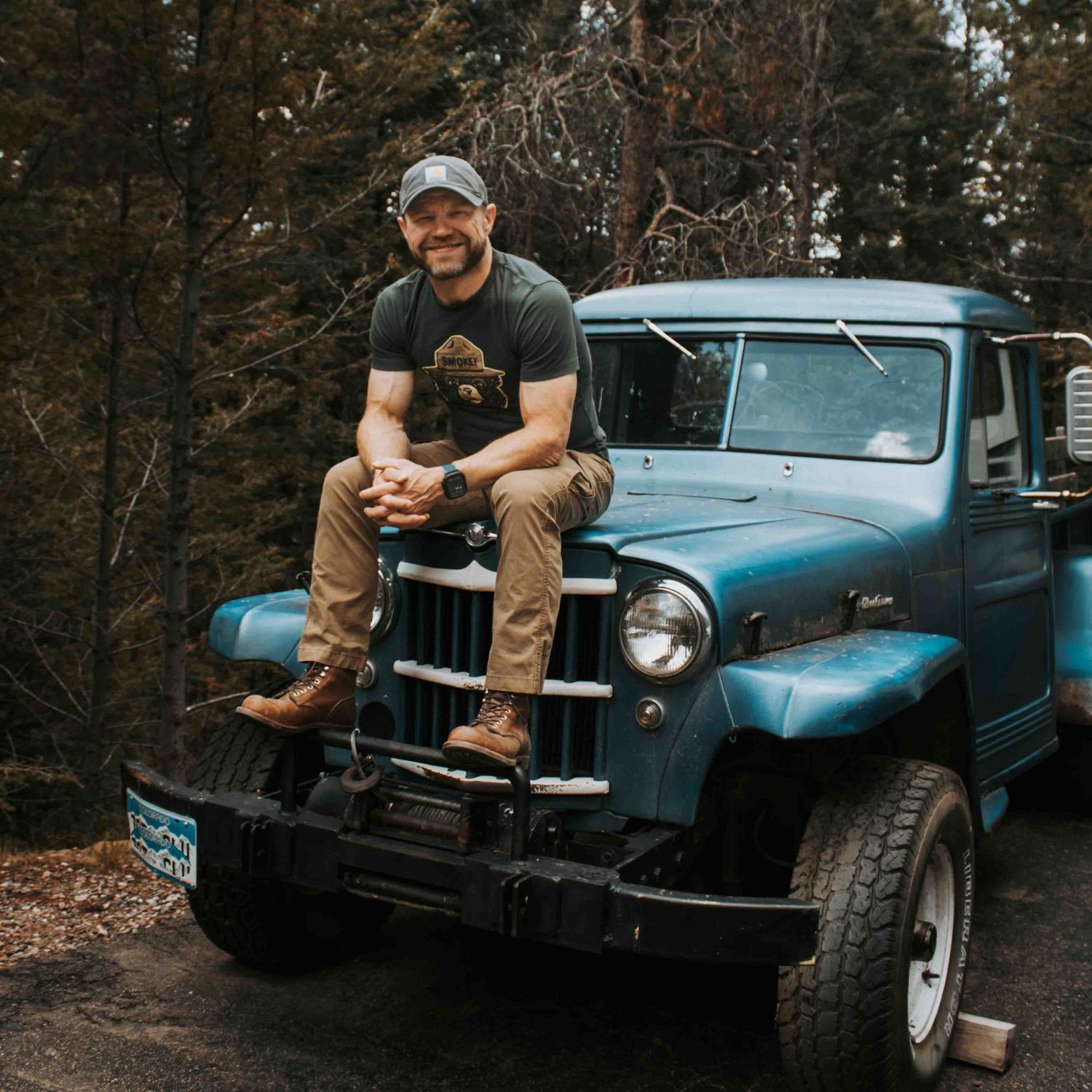 Man in cap and t-shirt sitting on hood of classic blue Willys pickup truck surrounded by pine trees.