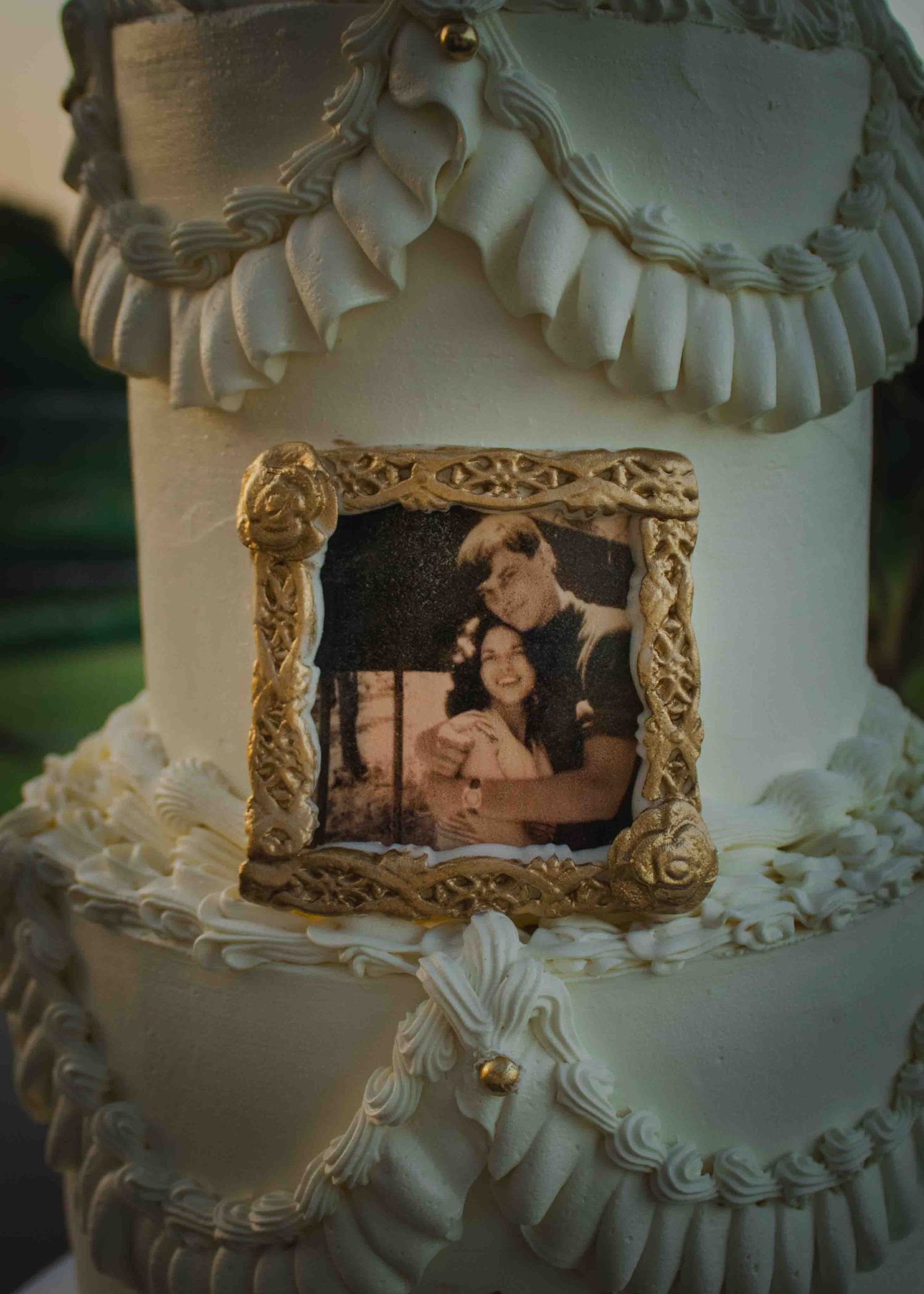 Elegant white wedding cake with gold-framed photo of couple, decorated with fondant ruffles and pearl accents.