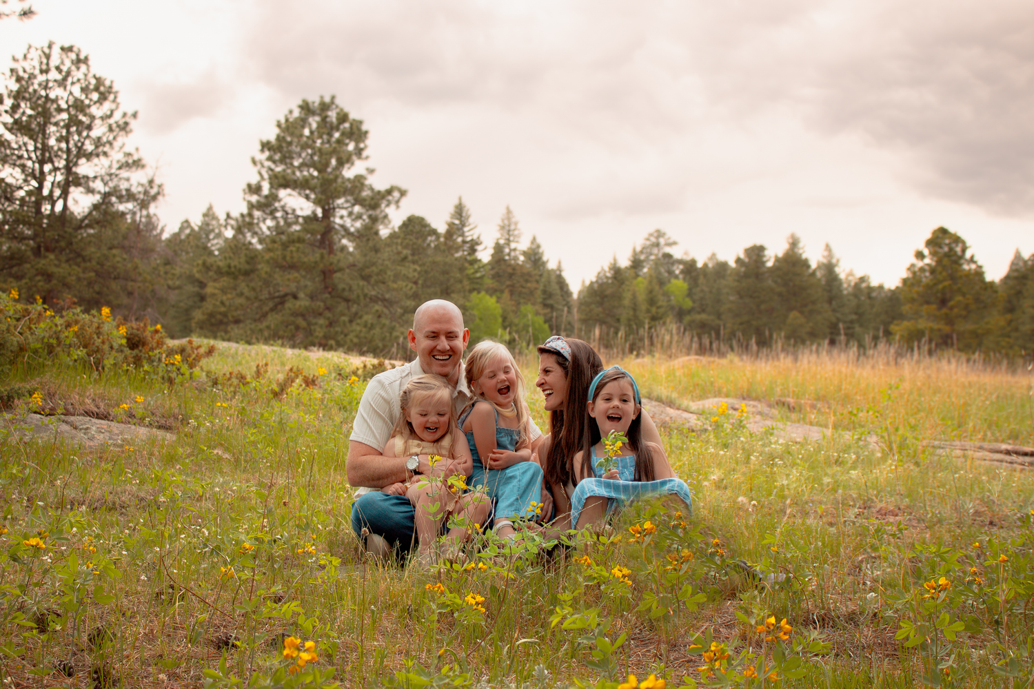 Happy family of five posing in a sunny wildflower meadow with pine trees in the background.