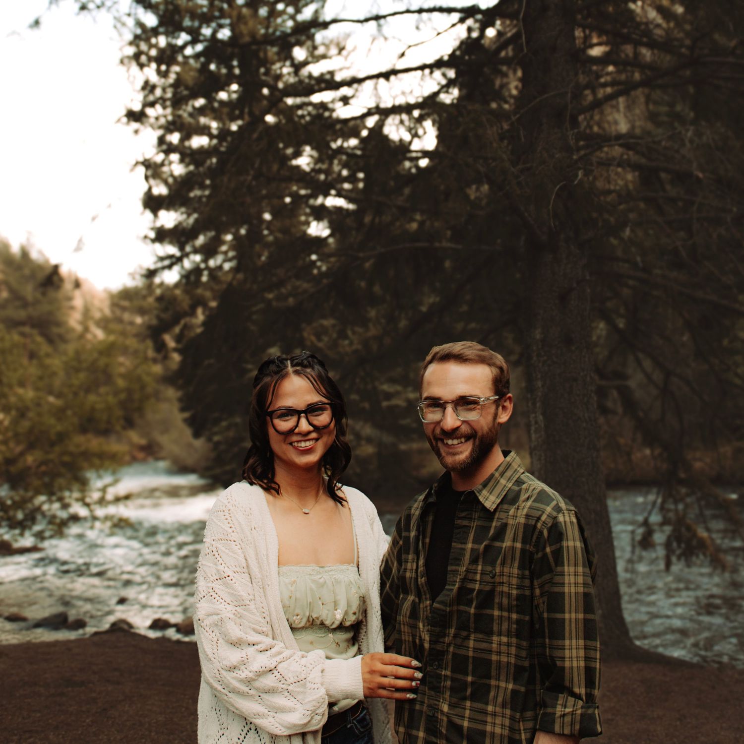 Couple smiling together outdoors near a river surrounded by tall pine trees in a forest setting.