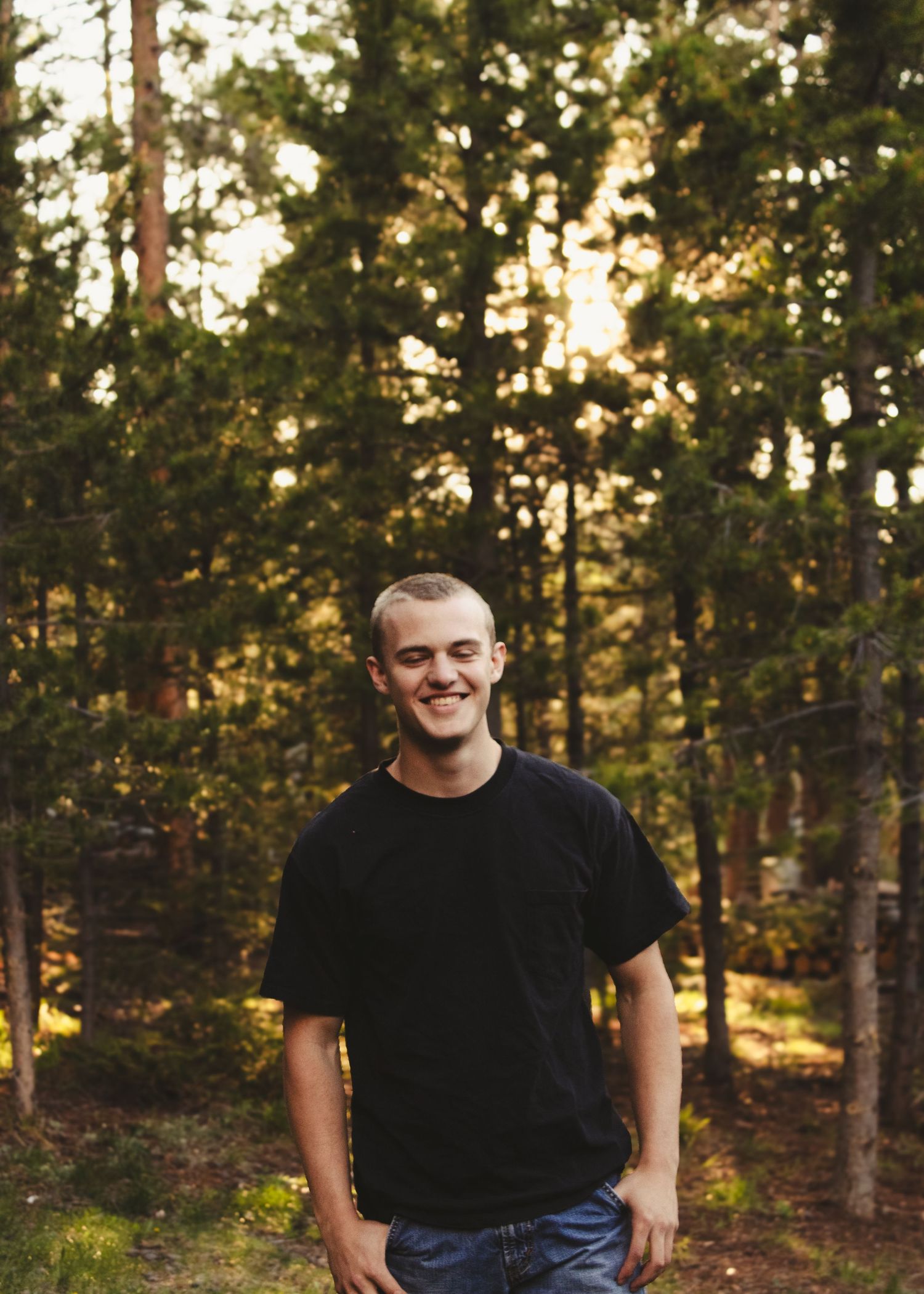 A smiling young man in a black t-shirt stands outdoors in a sunlit pine forest at golden hour.
