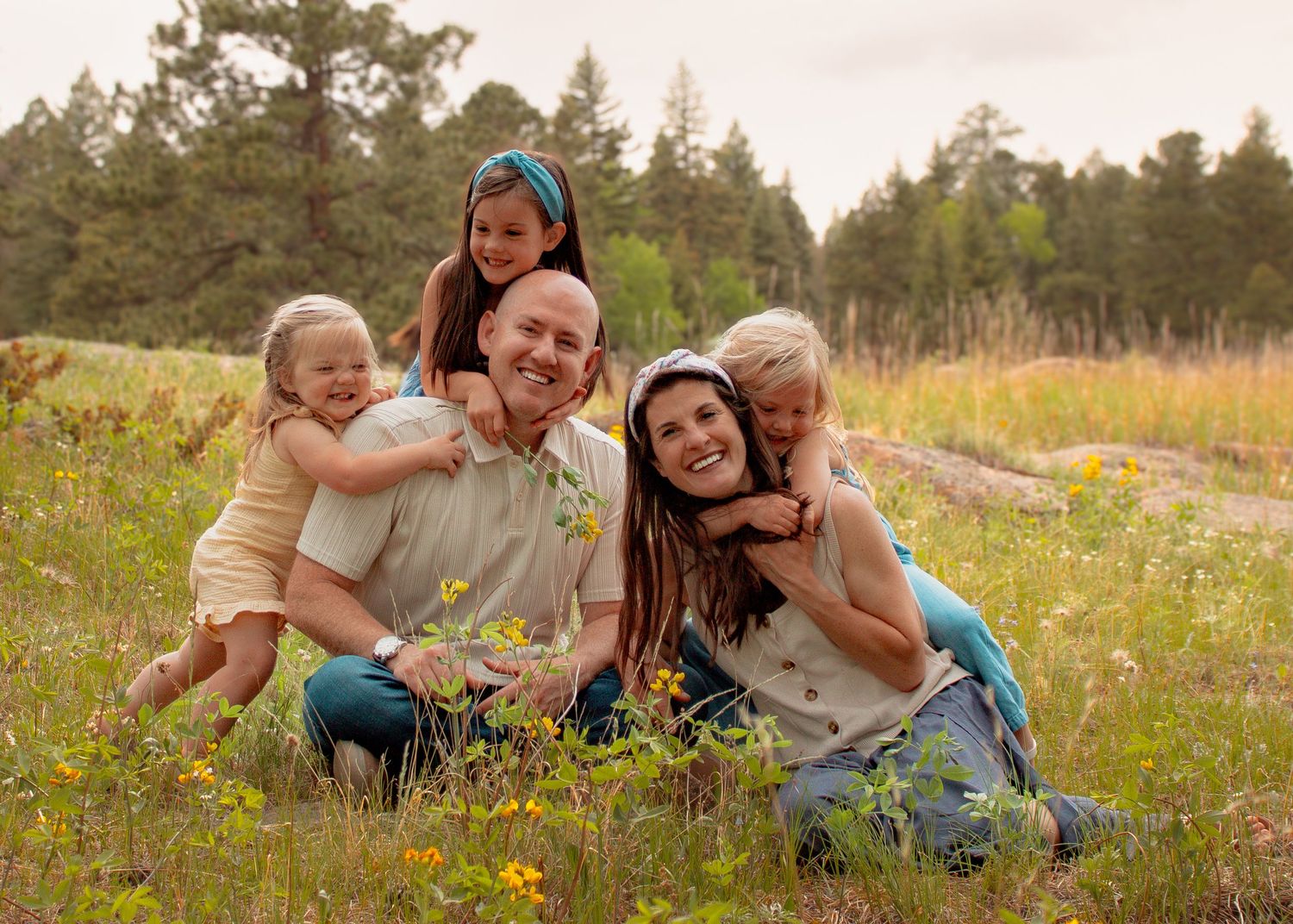 Happy family of five smiling together in a sunny wildflower meadow surrounded by pine trees.