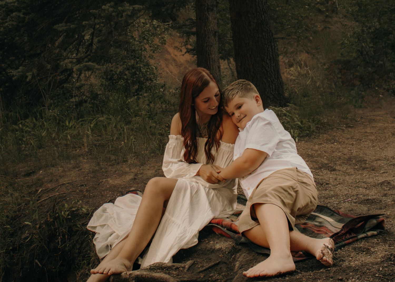 A mother in a white dress sits with her young son on a rock in a forest setting.