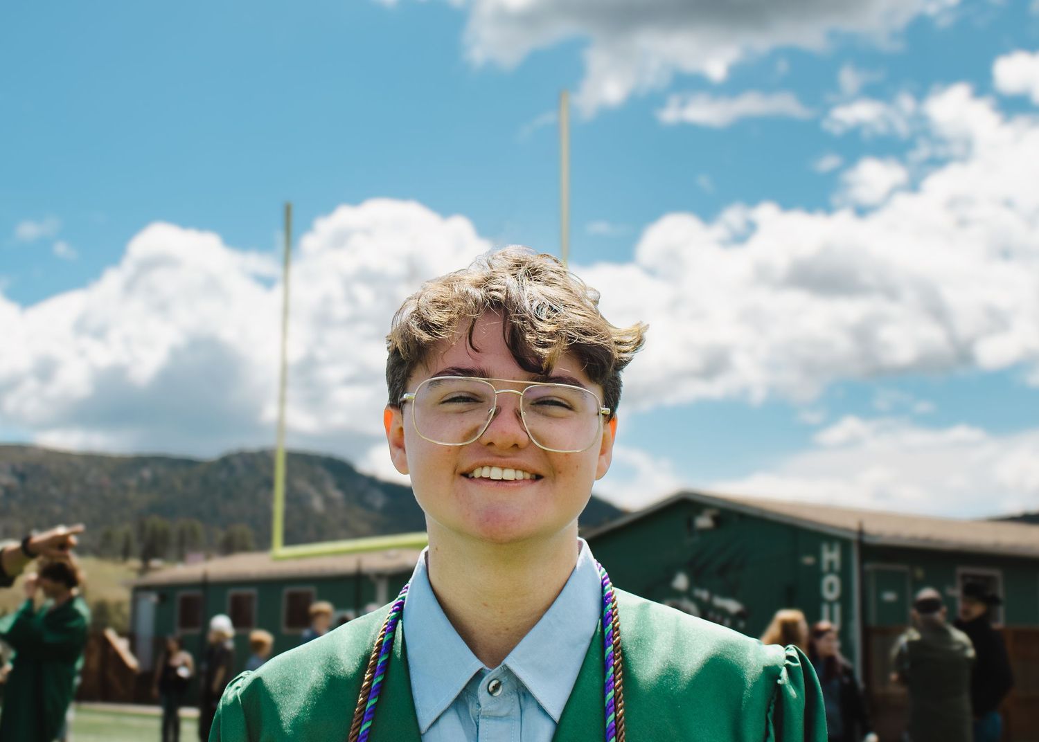 Smiling graduate in green gown and glasses celebrating outdoors on a sunny graduation day.