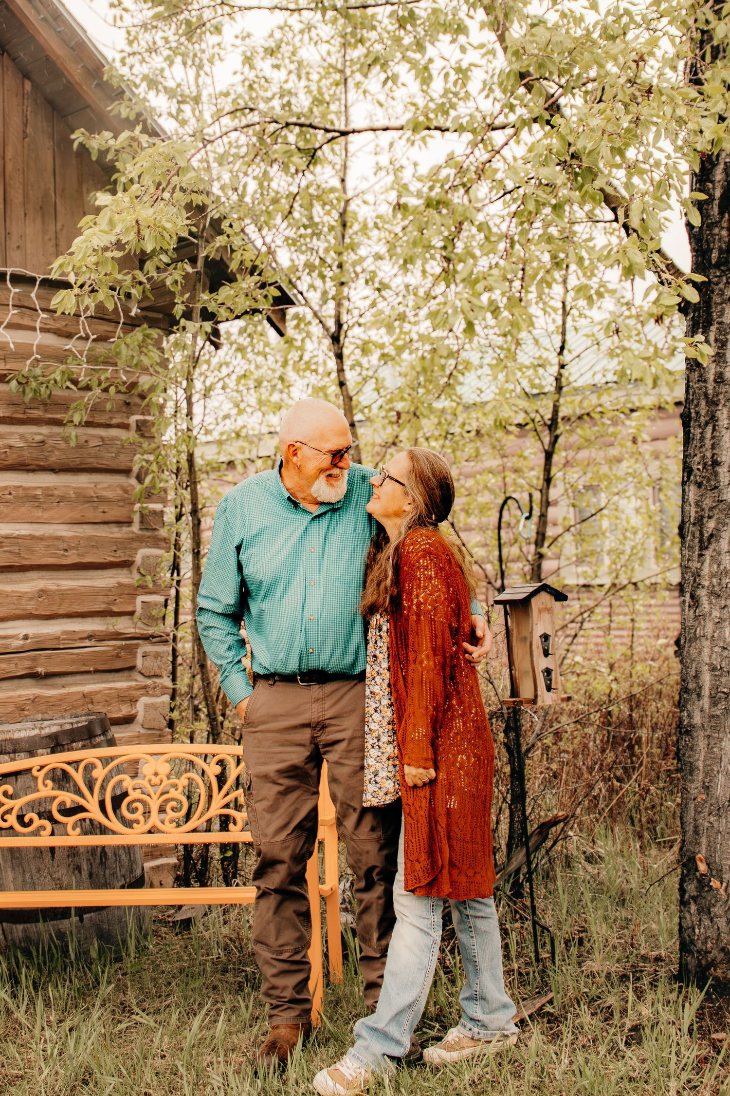 Elderly couple sharing a tender moment near a log cabin with a decorative bench and birdhouse.